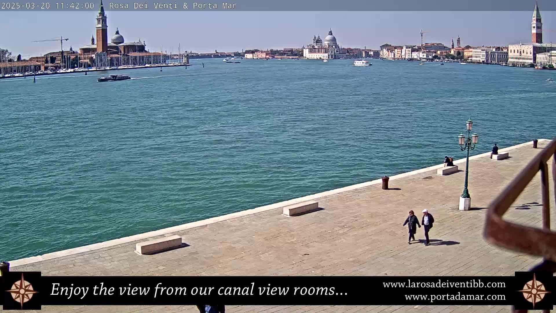 A sunny day in Venice shows a couple walking on a waterfront paved area overlooking a canal with boats and the distant city skyline including several prominent domes and bell towers.