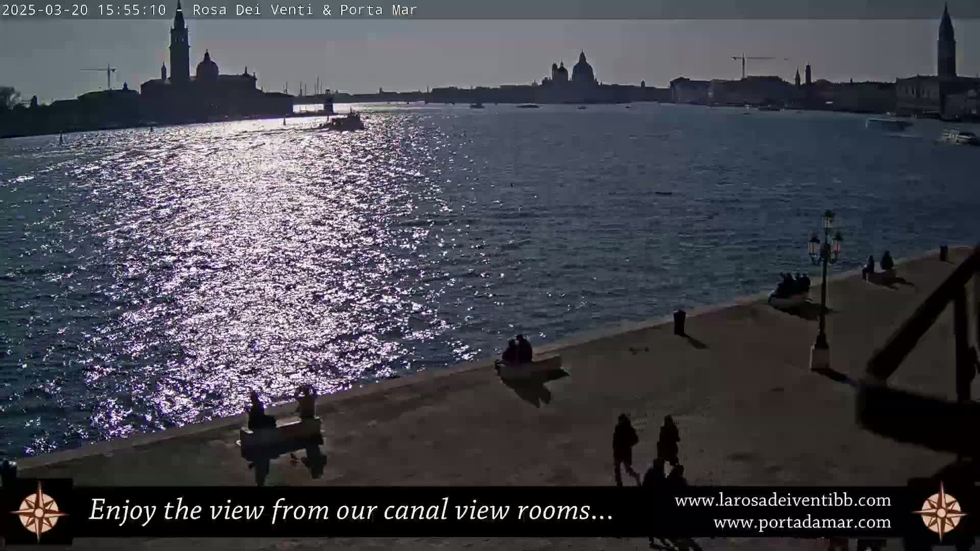 A sunlit canal scene shows a cityscape across the water with several people walking along a waterfront path.