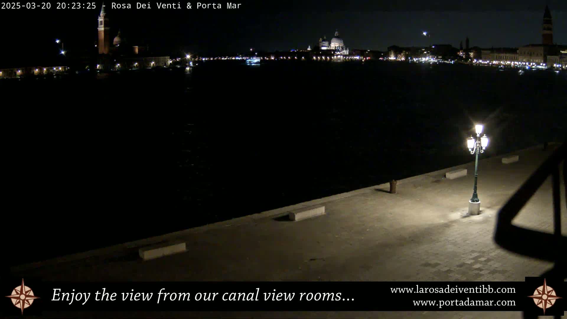 A nighttime view of a dark canal, with city lights and buildings visible in the distance, is seen from a waterfront walkway illuminated by a single lamppost.