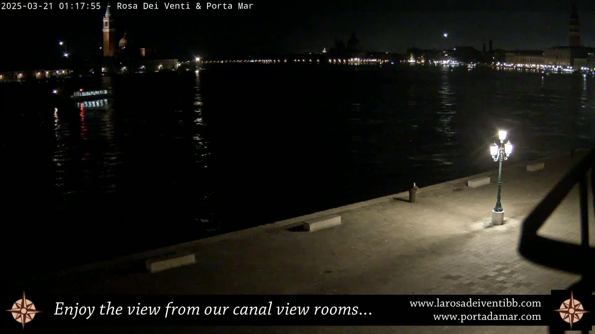 A nighttime view of a calm canal reflecting city lights, with a single lamppost illuminating a paved waterfront area.