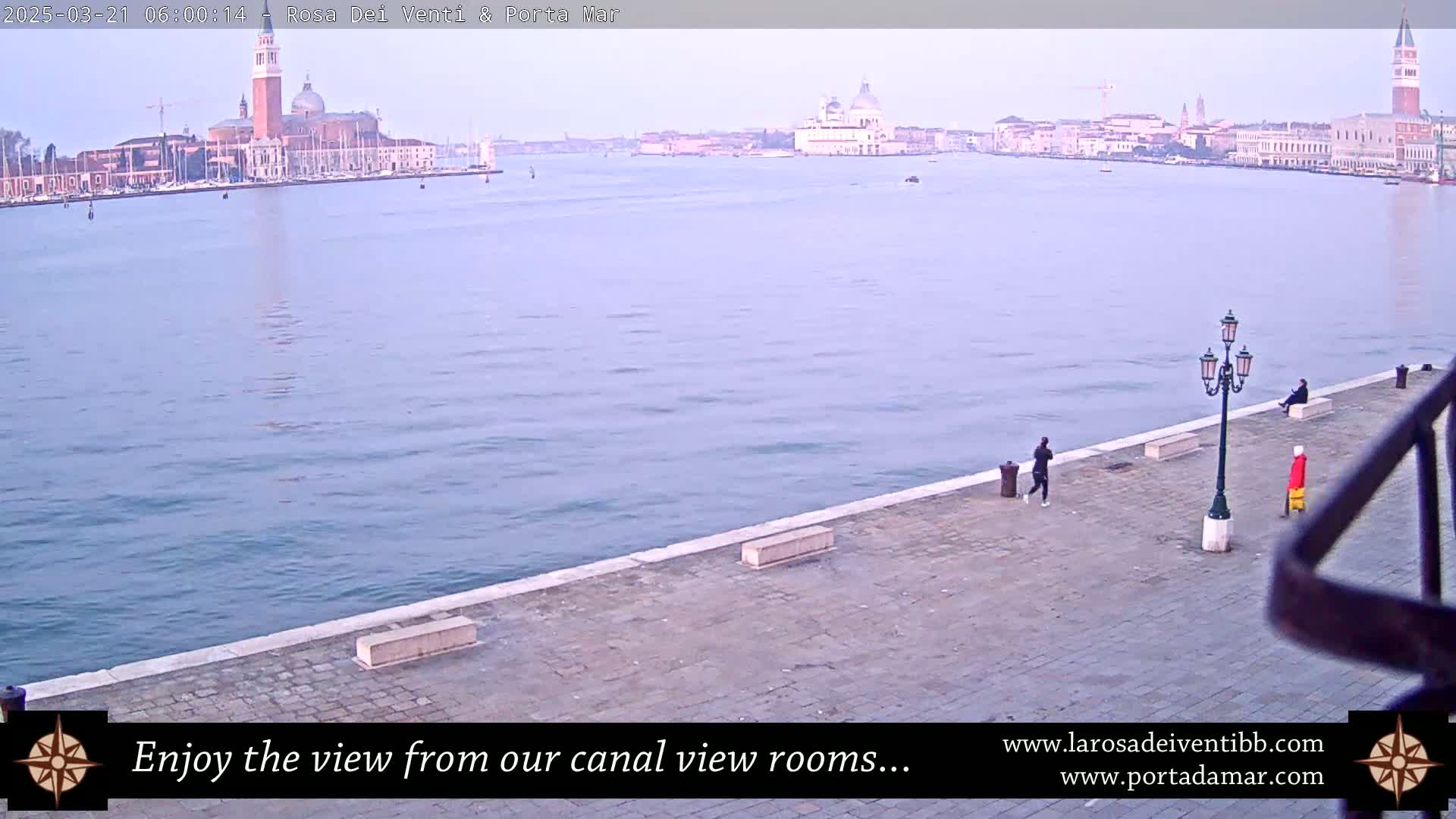 A calm, pale-blue canal stretches towards a cityscape of Venetian buildings under a clear, dawn sky, with a few people strolling along a paved waterfront.