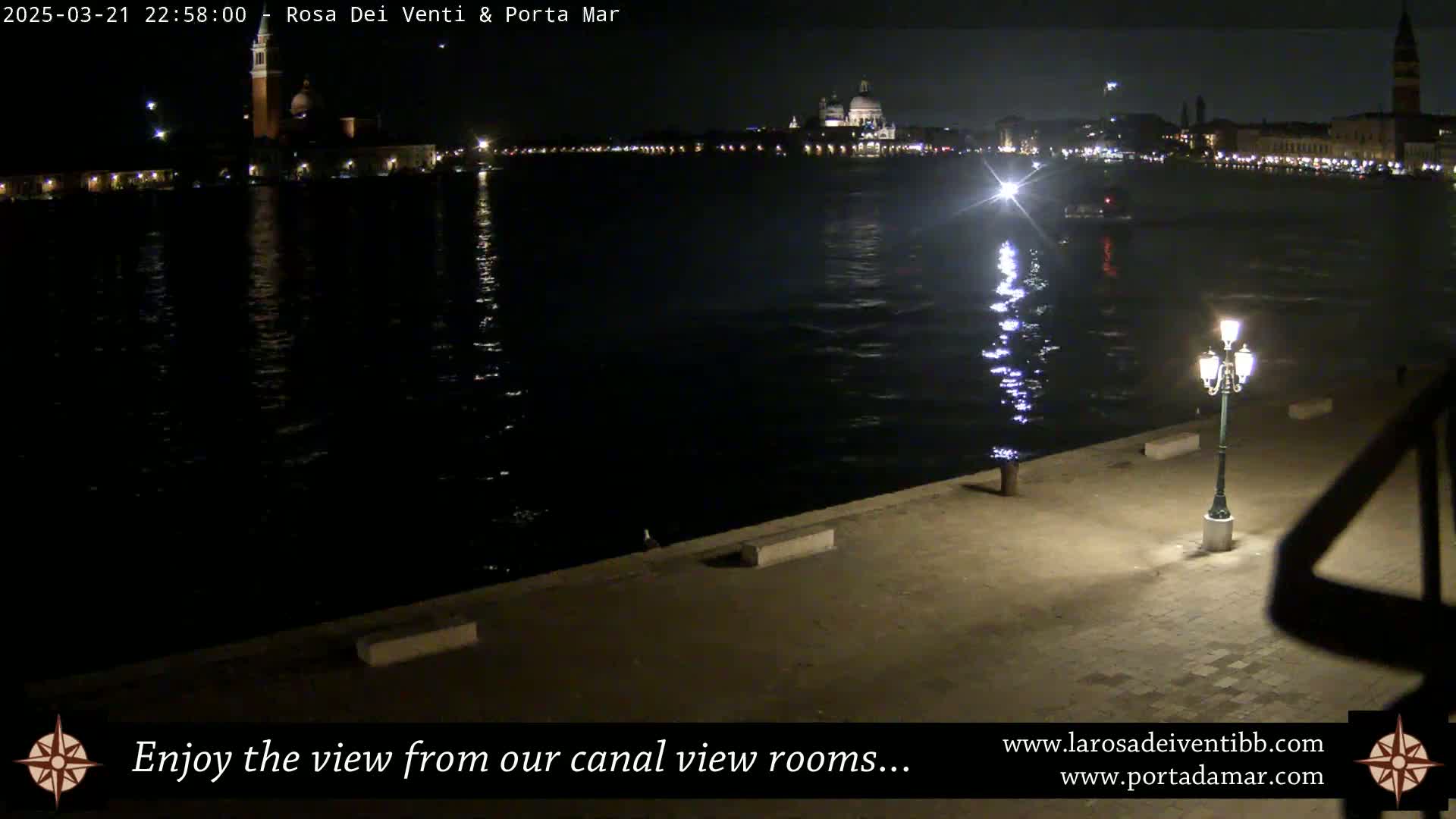 A nighttime view of a calm canal reflecting the lights of a city across the water, with a single lamp illuminating a paved waterfront area.