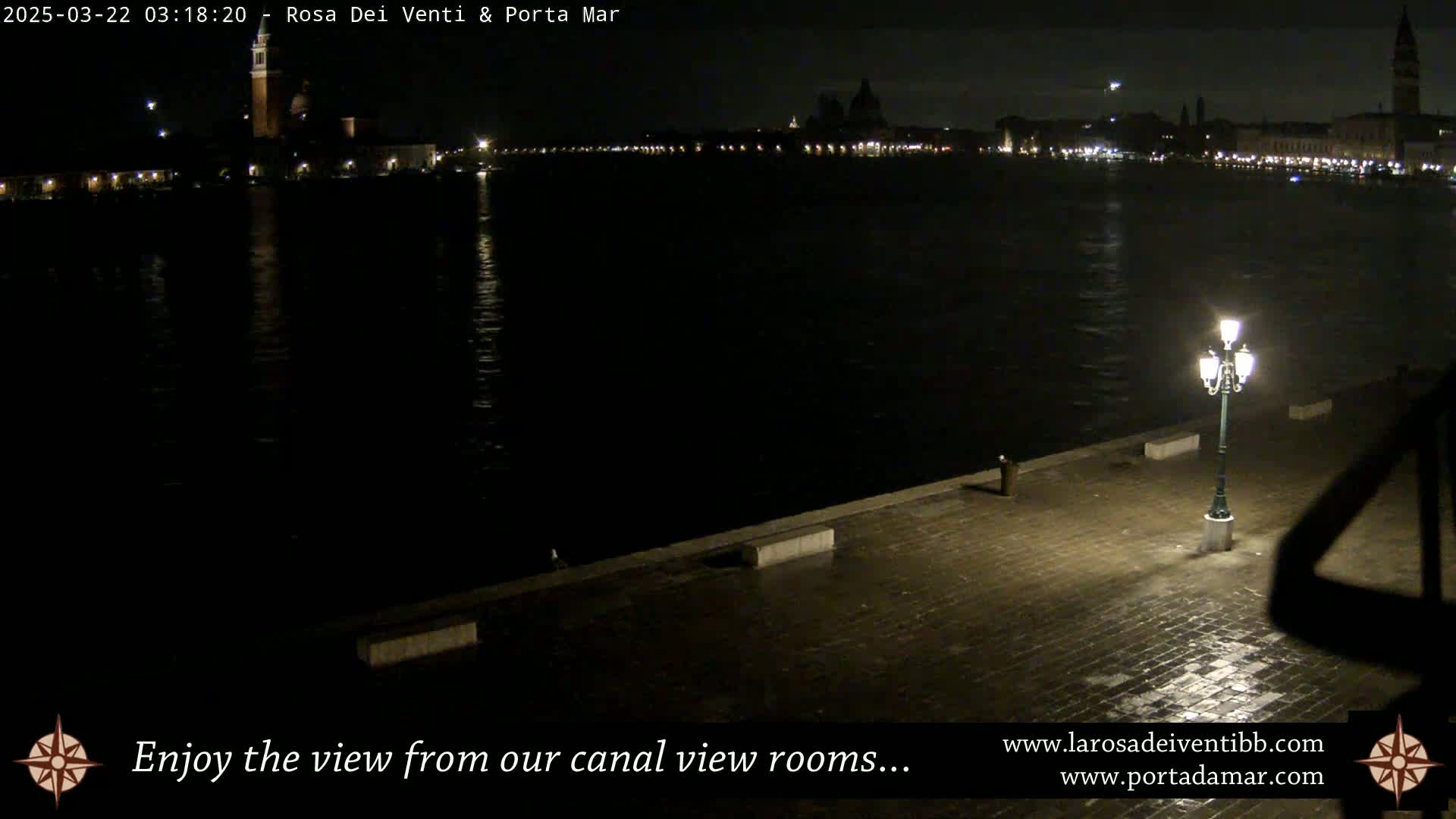 A nighttime view of a calm canal reflecting the lights of a distant city, seen from a paved waterfront with a single lit lamppost.