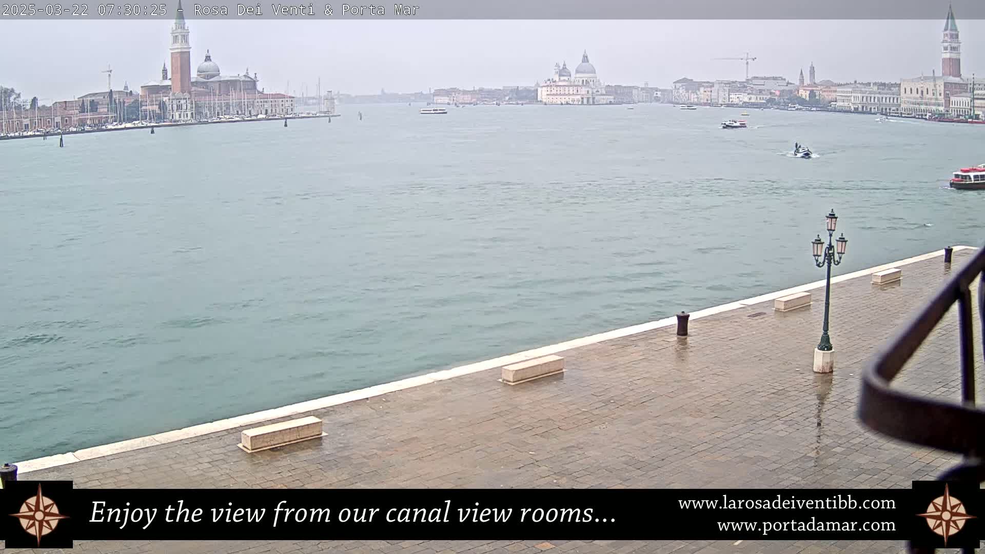 A paved waterfront area shows a view of a calm canal with distant buildings and boats under an overcast sky.