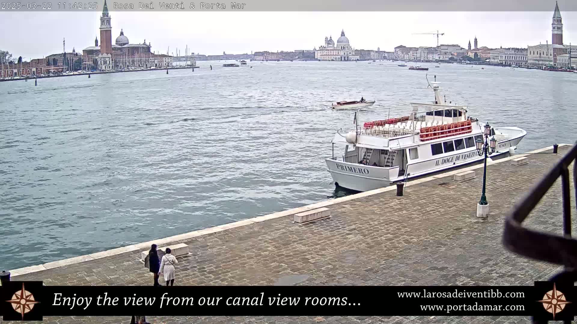 A large passenger boat is docked at a waterfront paved area, with two people standing nearby, overlooking a calm canal with a distant view of Venetian architecture under an overcast sky.