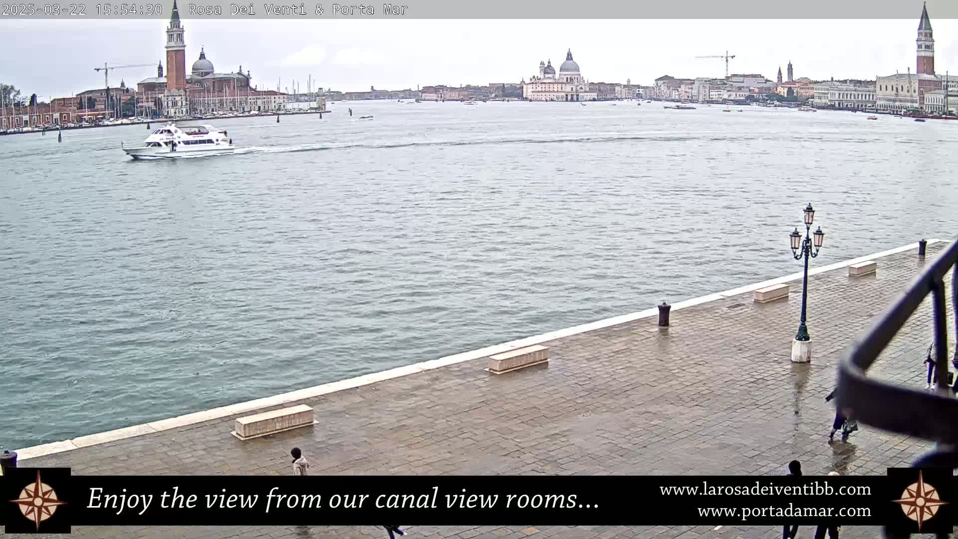 A waterfront paved area with a few people walking, overlooking a calm canal with a boat and the distant cityscape of Venice under an overcast sky.