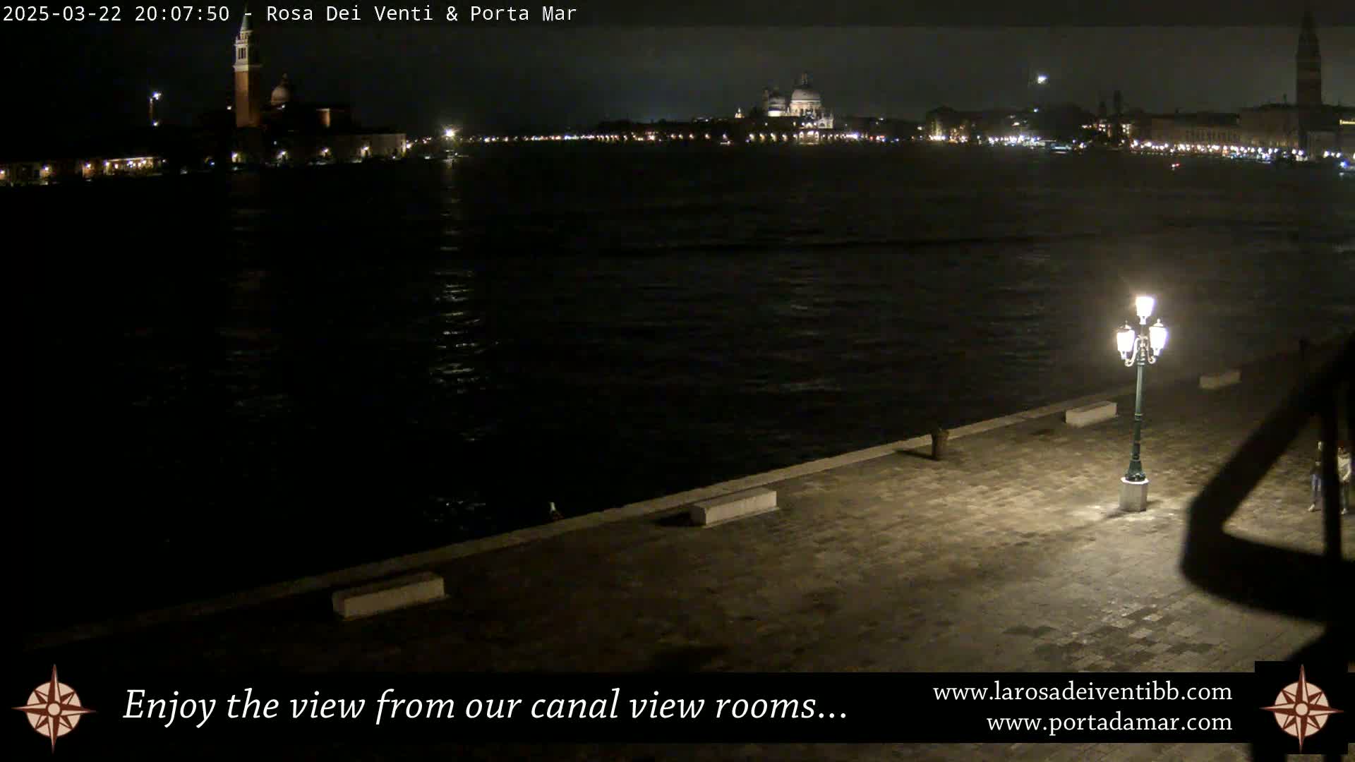 A nighttime view across a dark canal shows illuminated buildings on the far shore and a single lit lamppost on a paved walkway in the foreground.