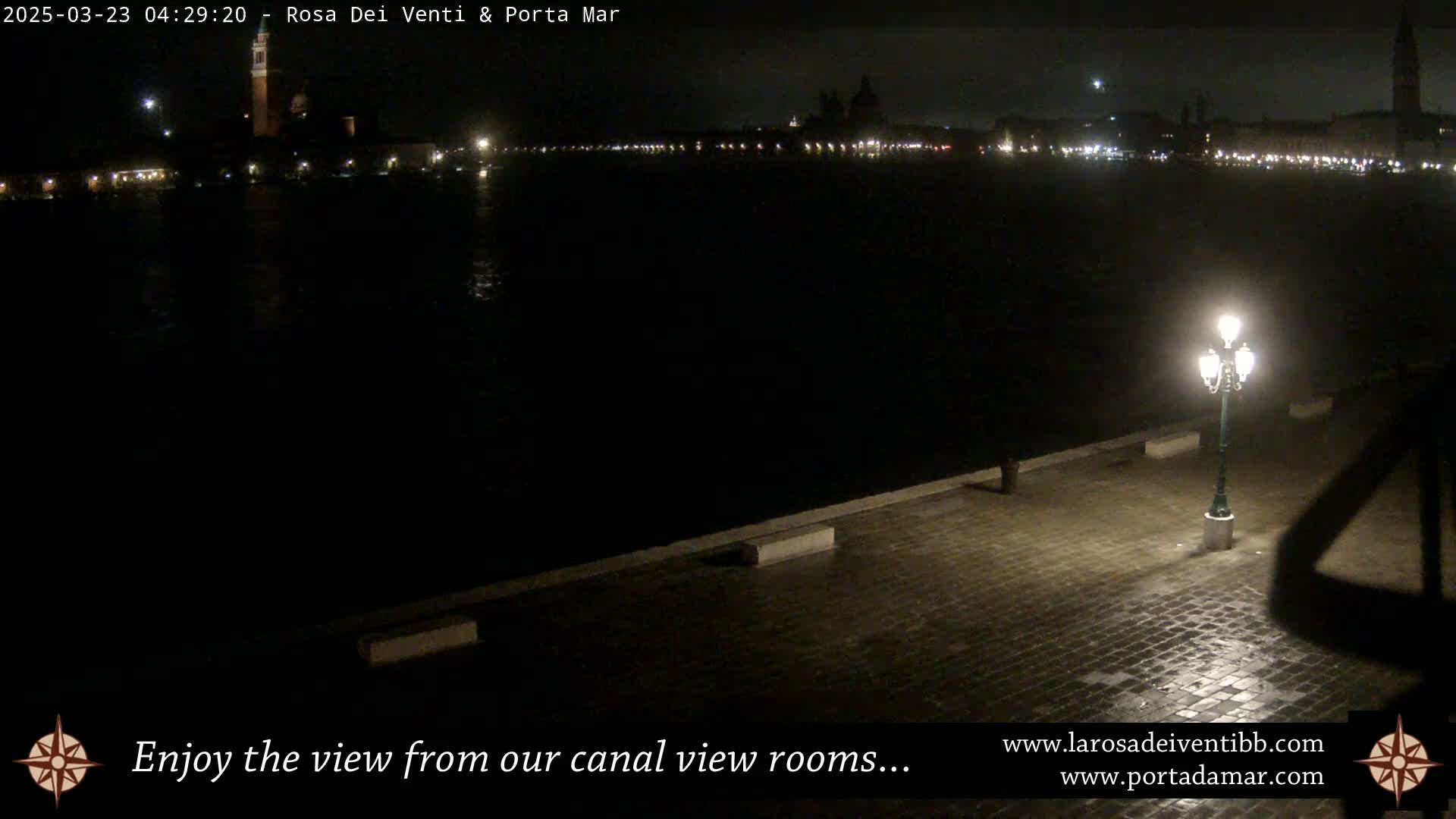 A nighttime view of a city across a dark body of water, illuminated by distant lights and a single lamppost on a paved waterfront.