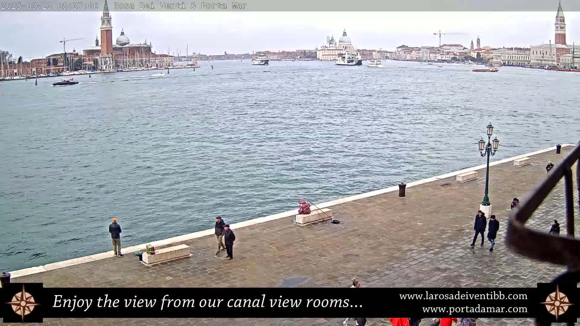 A waterfront view of a calm canal in Venice, with several people walking along a paved embankment and a distant cityscape featuring prominent church domes under an overcast sky.
