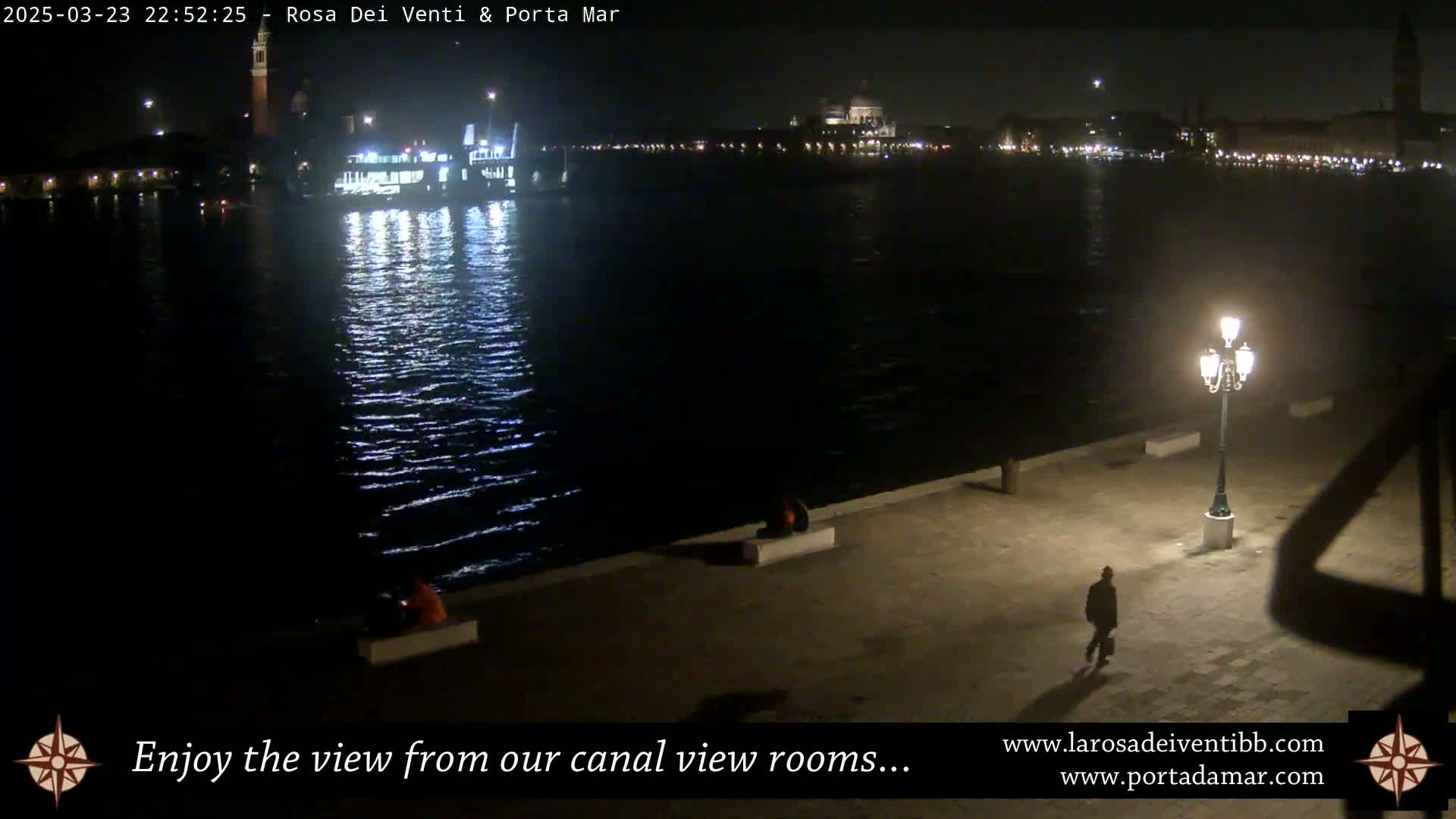 A nighttime view of a canal with a boat, city lights in the distance, and a lone person walking along a waterfront walkway under a streetlight.