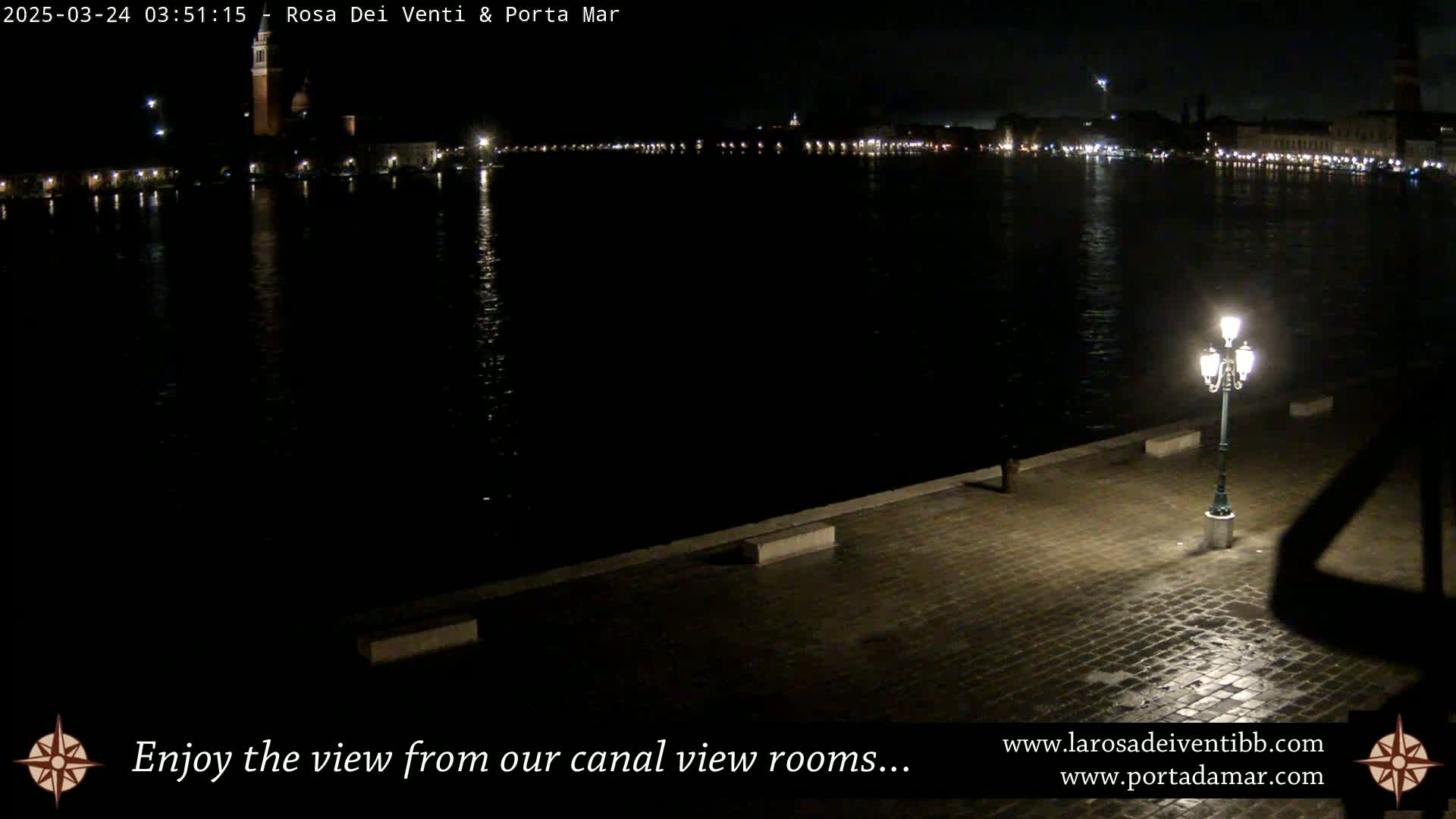 A nighttime view across a dark canal shows a city skyline illuminated with lights, and a single lit lamppost on a cobblestone waterfront.