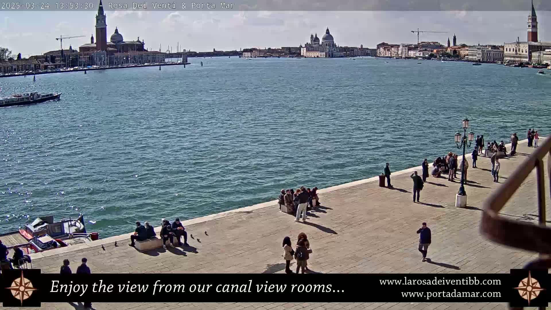 A sunny day in Venice shows a wide canal with buildings and a church in the distance, and several people sitting and walking along a paved waterfront.