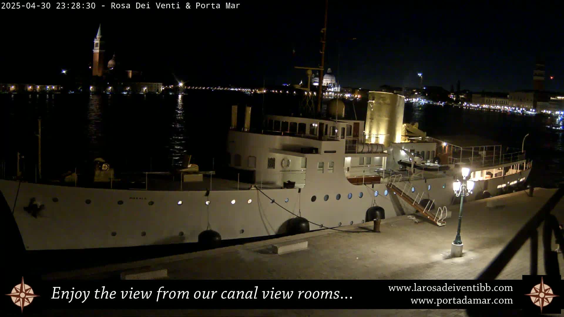 A large, white ship is docked at night, with a city skyline illuminated across the water.