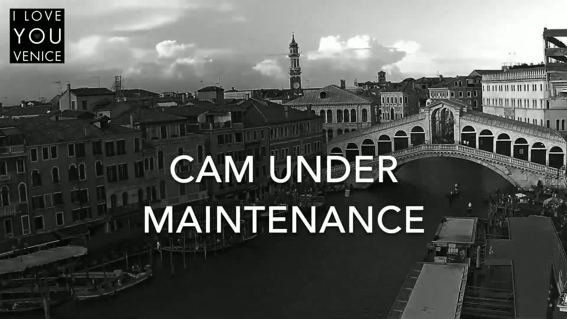 A black and white, high-angle view shows a canal in Venice, Italy, under a partly cloudy sky, with a bridge and buildings lining the waterway.