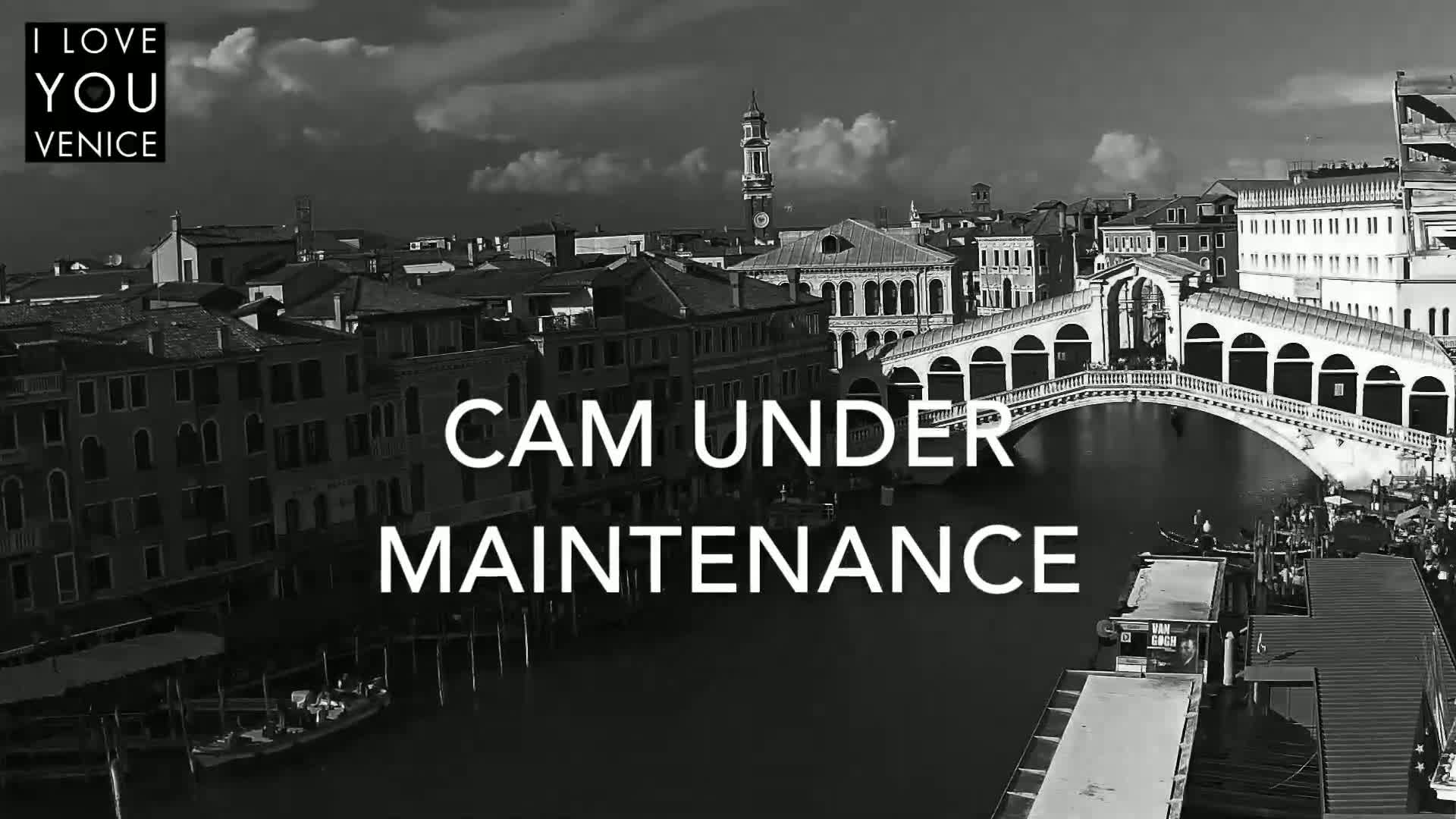 A black and white, high-angle view shows a Venetian canal with boats, buildings, and the Rialto Bridge under a partly cloudy sky.