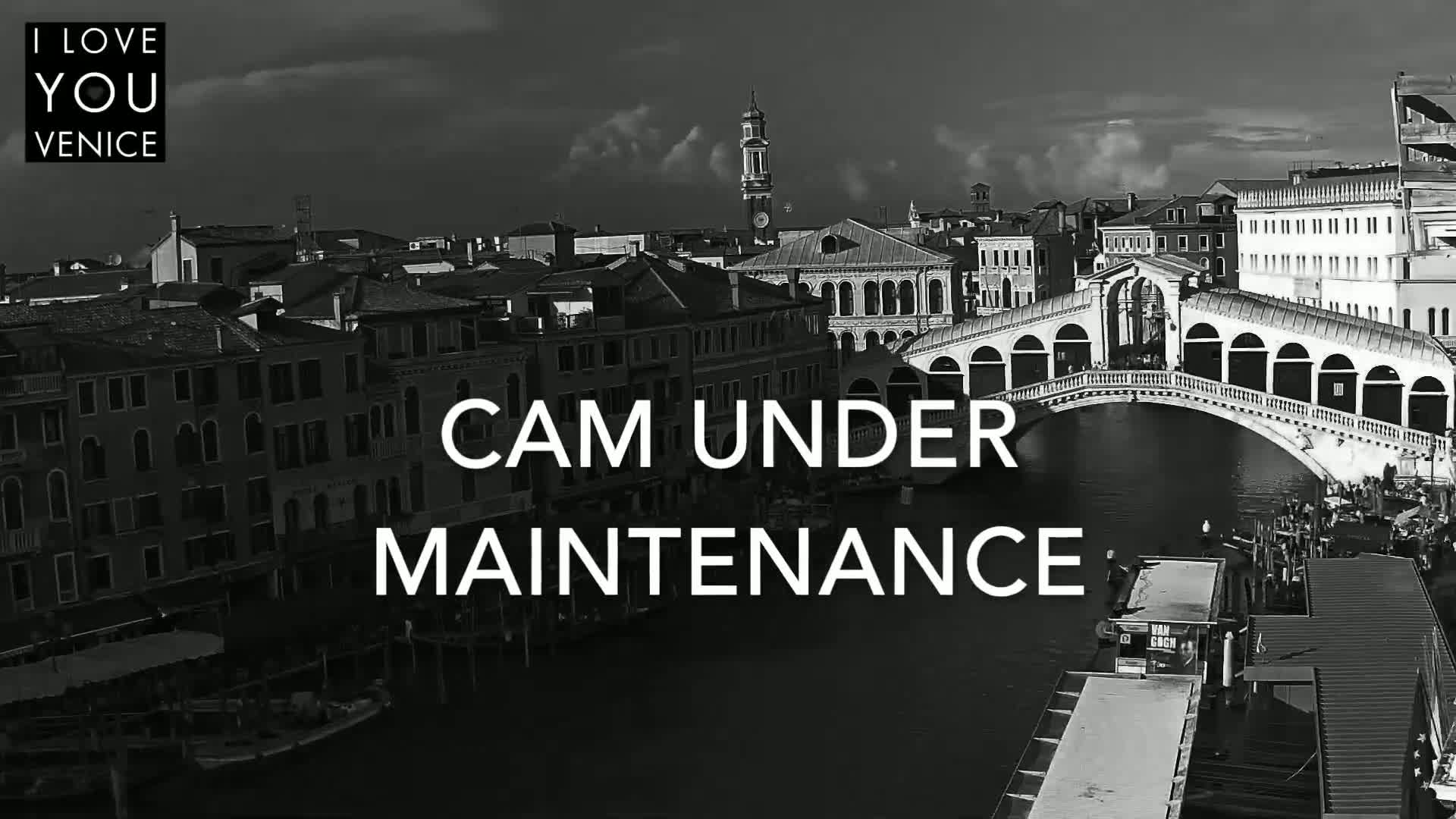 A black and white, high-angle view shows a canal in Venice with a bridge and buildings lining the waterway under a partly cloudy sky.