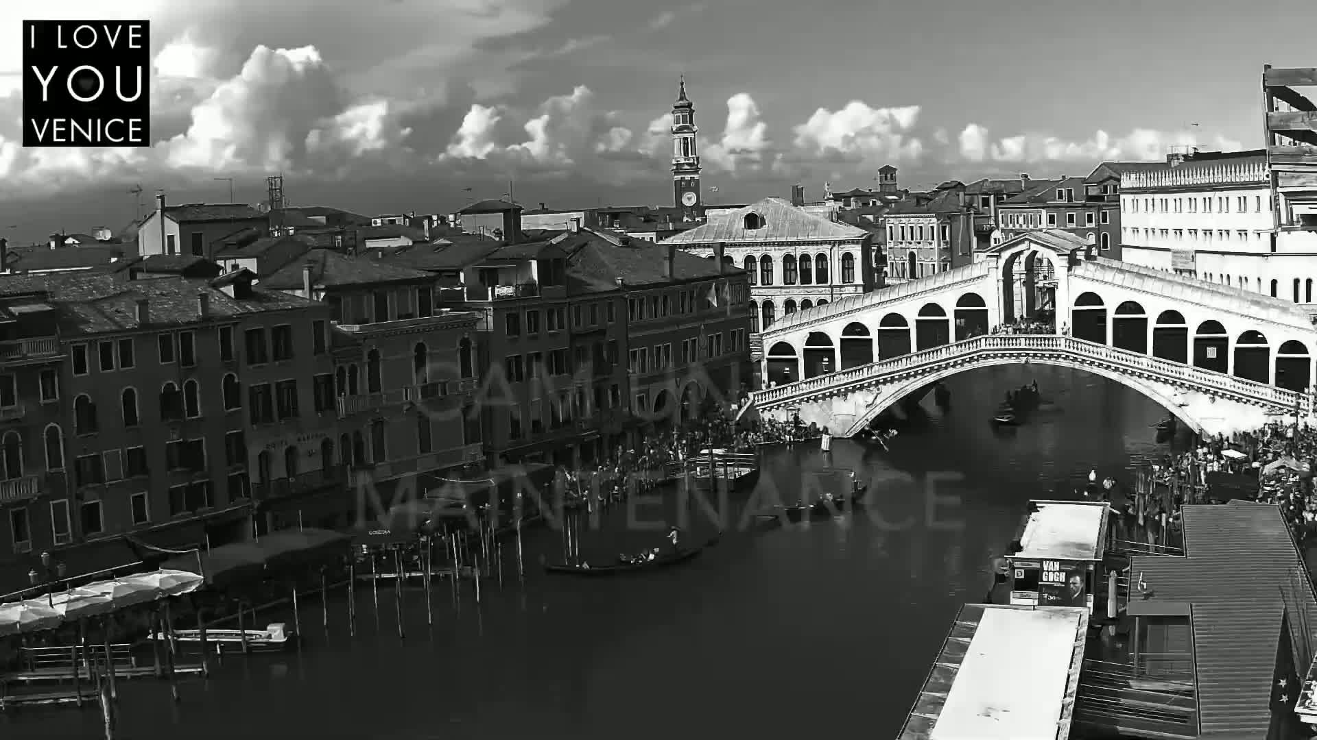 A black and white view of a bridge over a canal in Venice, under a partly cloudy sky, with numerous boats and people visible.