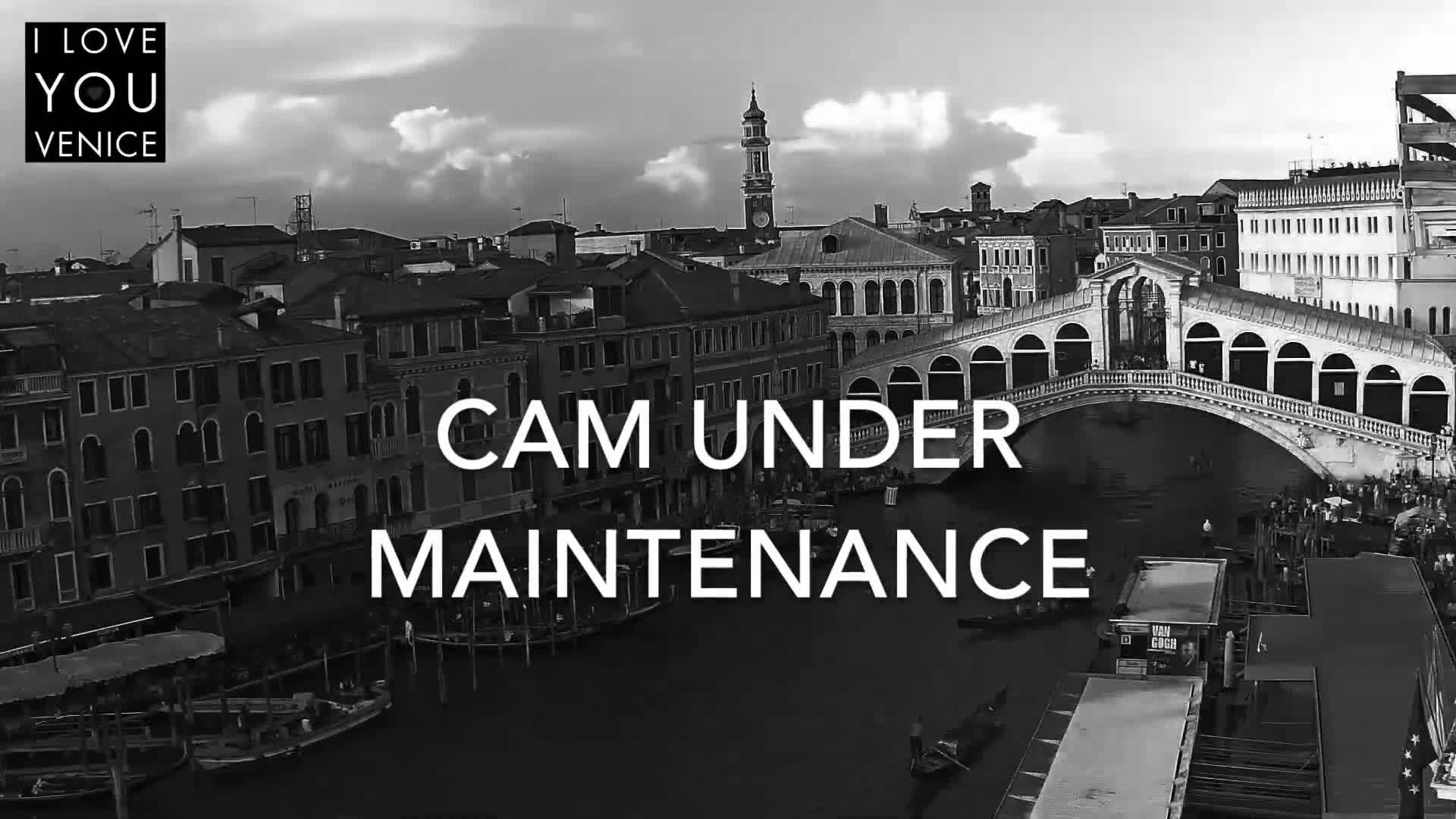 A black and white, high-angle view shows a canal in Venice, with a bridge arching over the water, boats moored along the banks, and buildings lining the waterway under a partly cloudy sky.