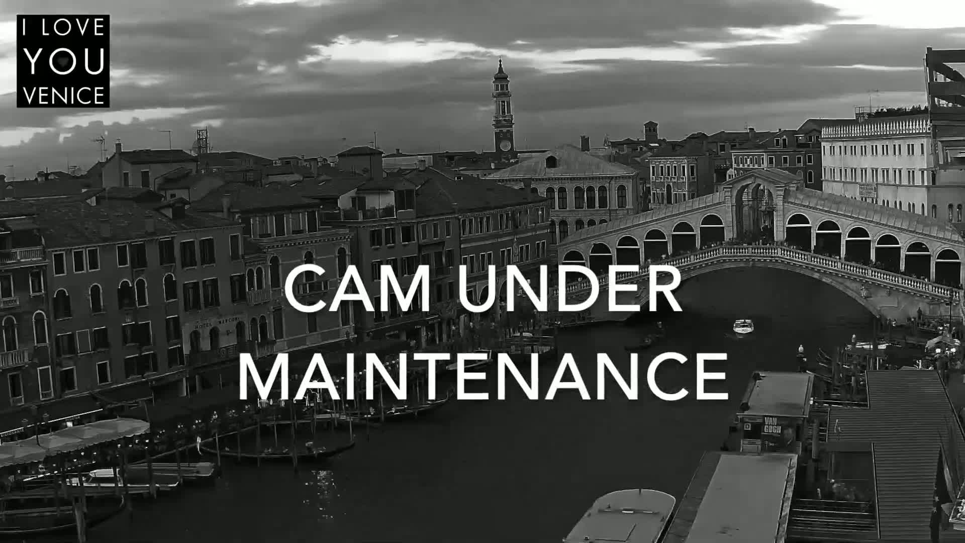 A black and white, high-angle view of a canal in Venice, Italy, shows boats moored along the waterway, buildings lining the canals, and a bridge spanning the water under cloudy skies.