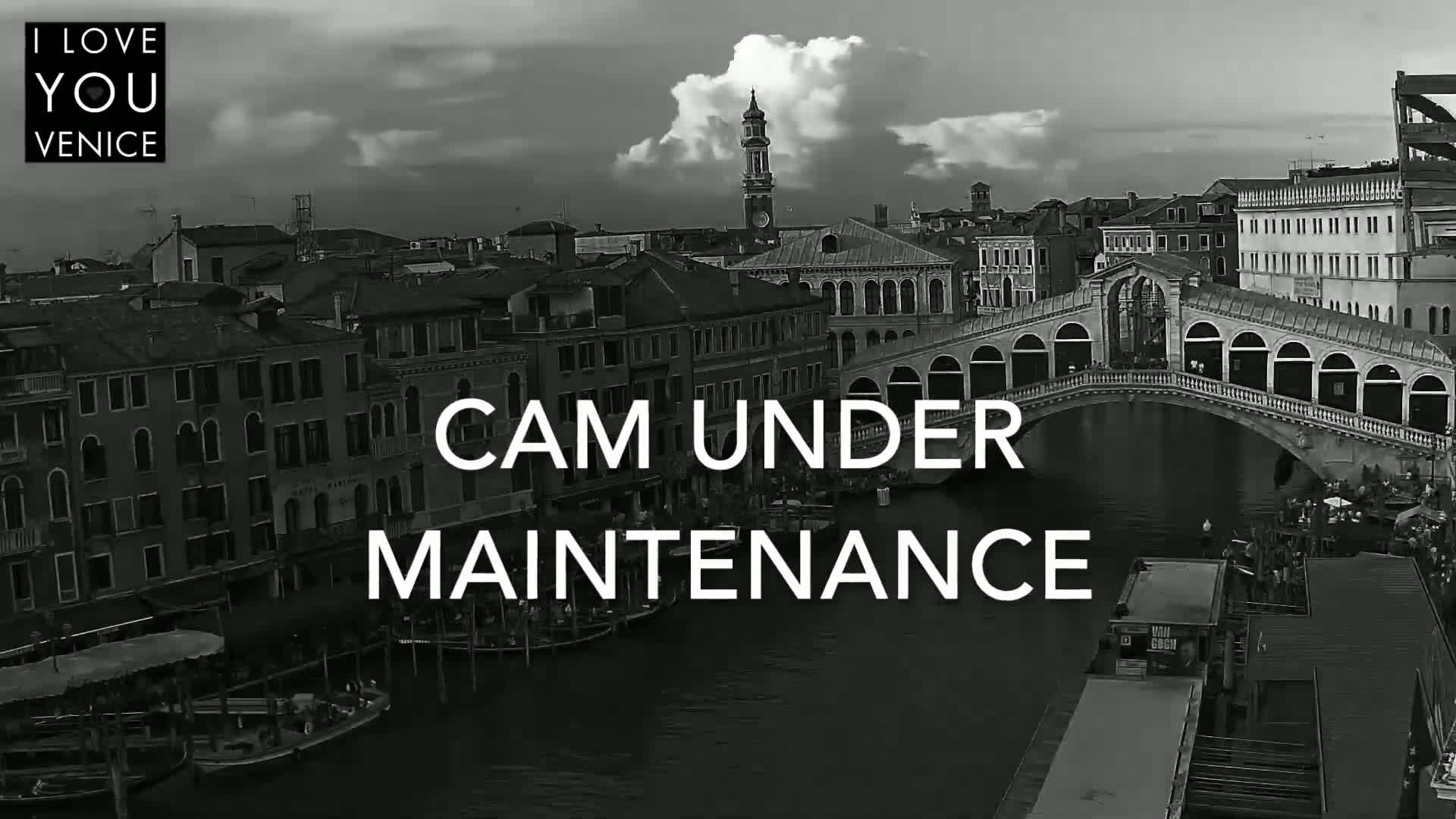 A black and white view of a Venetian canal, showing a bridge and buildings under a partly cloudy sky.