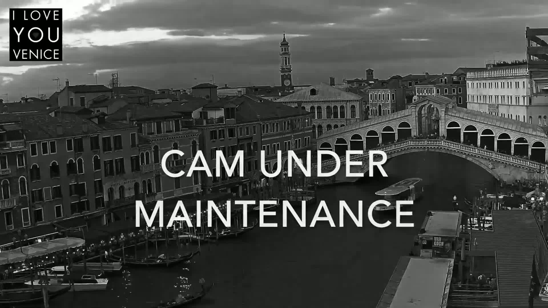 A grayscale view of a canal in Venice, under a cloudy sky, shows a bridge with boats and buildings along its sides.