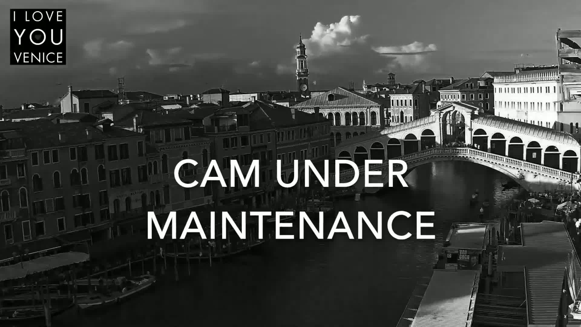 A black and white, high-angle view shows a bridge over a canal in Venice, with buildings lining the waterway under partly cloudy skies.