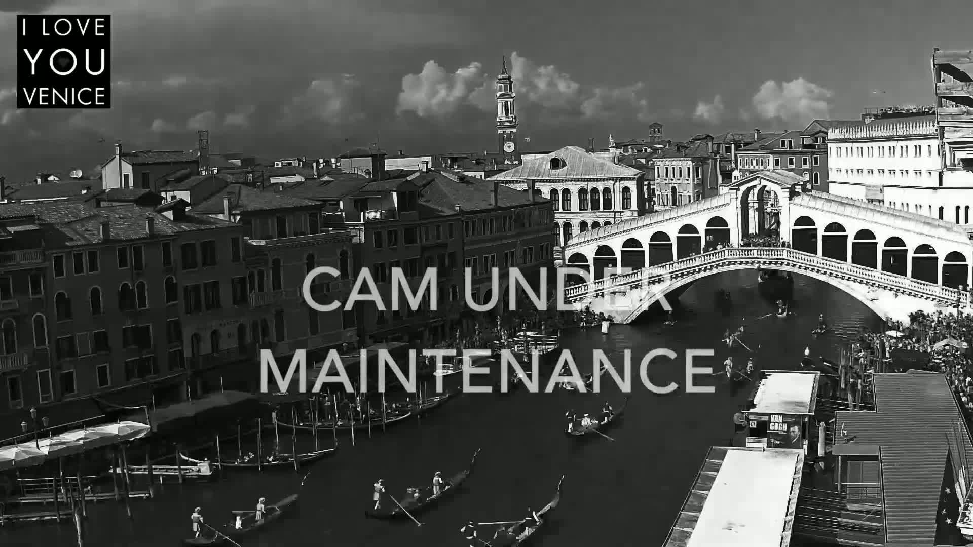 A black and white aerial view shows gondolas on a canal beneath a bridge in Venice, under a partly cloudy sky.