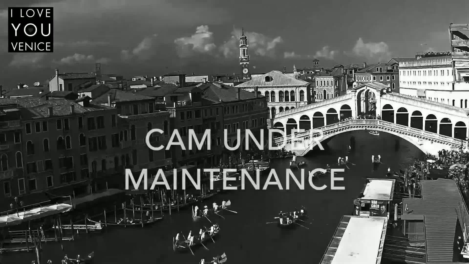 A black and white aerial view shows a canal in Venice, with gondolas on the water and the Rialto Bridge in the background under a partly cloudy sky.