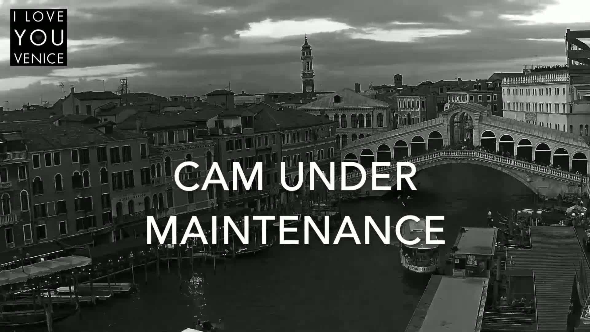 A black and white, high-angle view shows a Venetian canal at dusk with boats moored along the sides, buildings lining the canal, and the Rialto Bridge spanning the waterway.