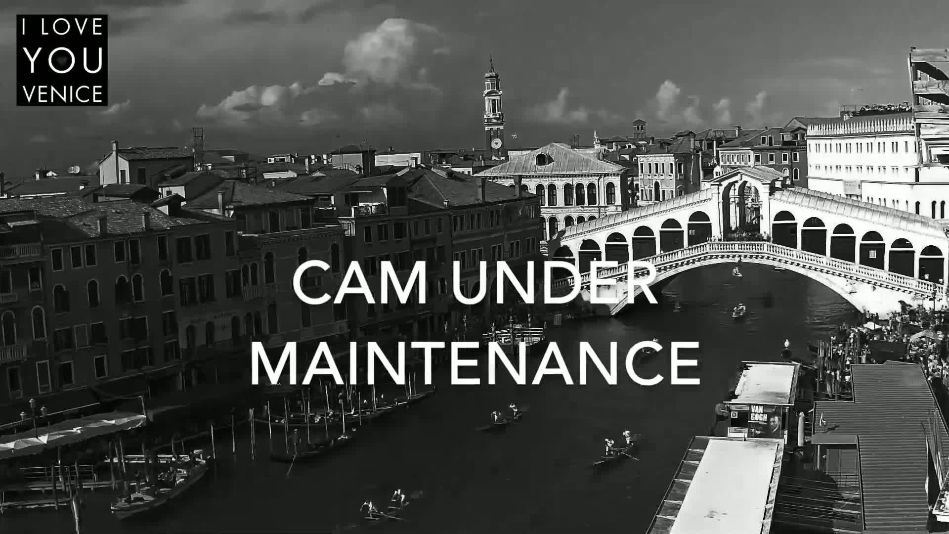 A black and white aerial view of a canal in Venice, Italy, shows a bridge with people walking across it and gondolas on the water under a partly cloudy sky.