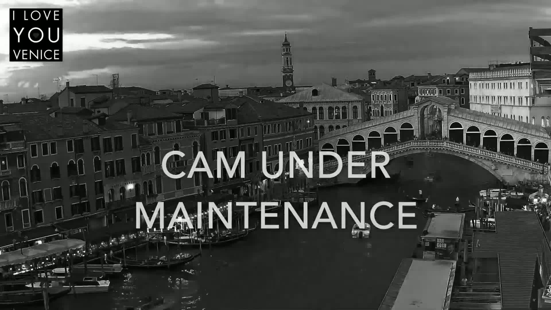 A black and white, nighttime view of a canal in Venice, showing gondolas, buildings, and a bridge under a partly cloudy sky.