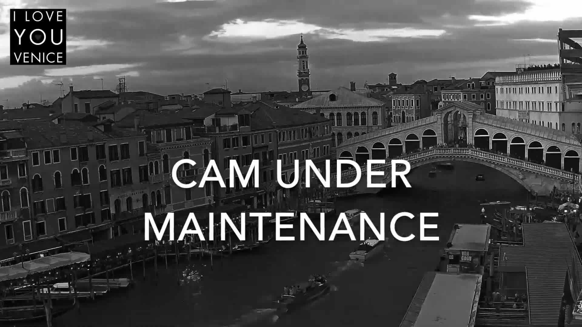 A black and white view of a Venetian canal, showing boats, buildings, and the Rialto Bridge under a cloudy sky.