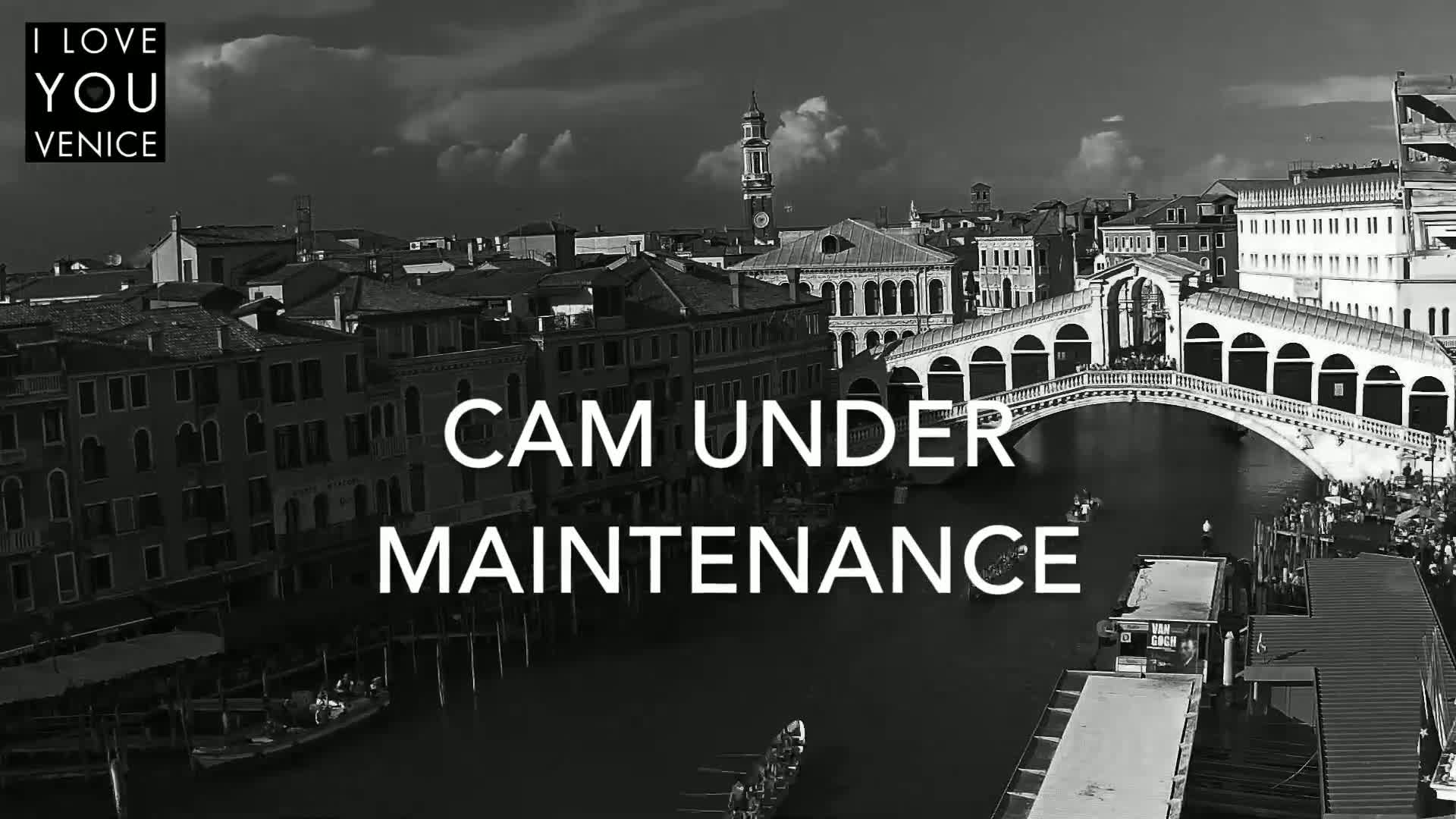 A black and white, high-angle view shows a canal in Venice, Italy, with gondolas, a bridge, and buildings under a partly cloudy sky.