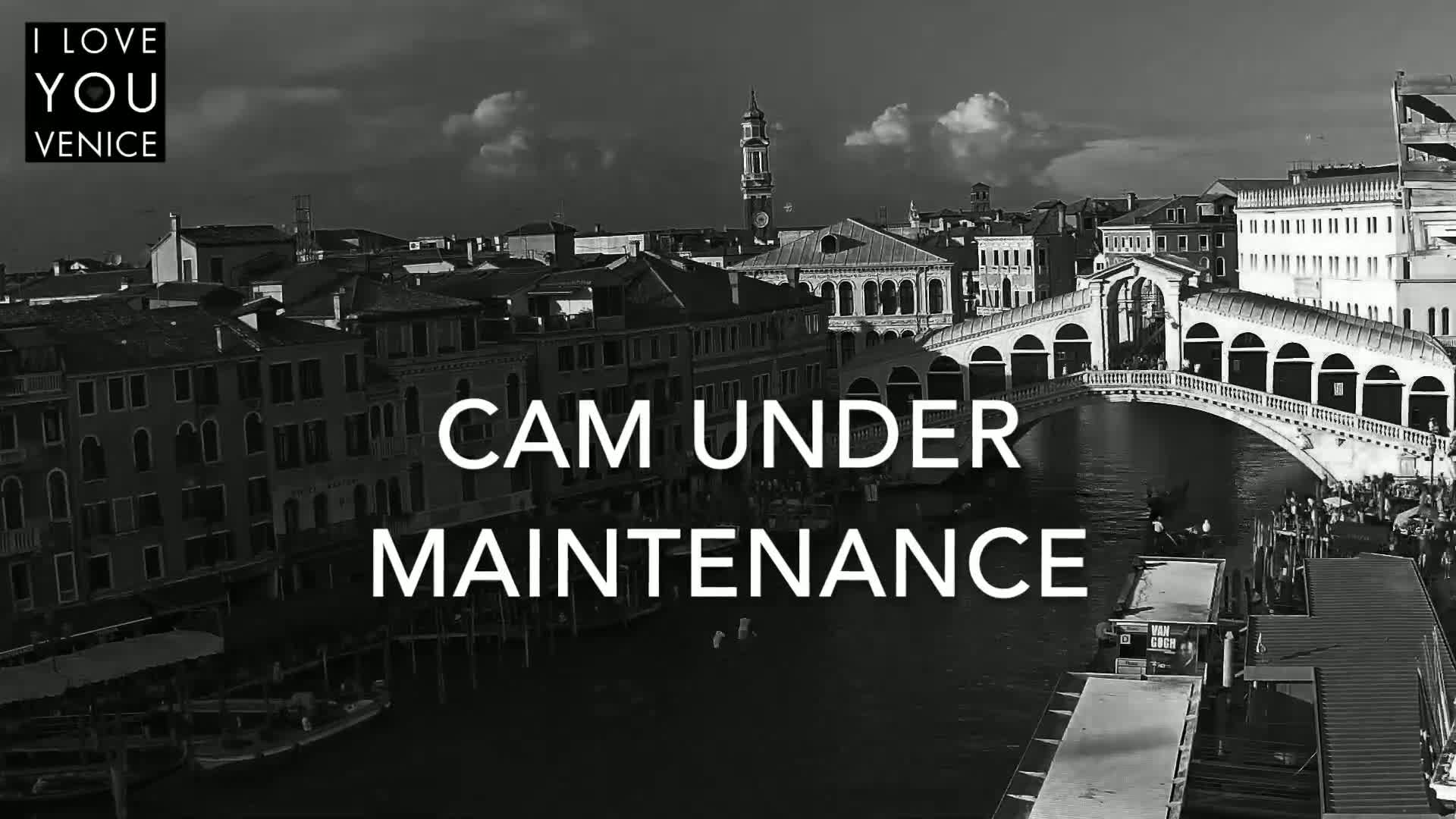 A black and white, high-angle view shows a canal in Venice, Italy, with a bridge and numerous buildings under a partly cloudy sky.