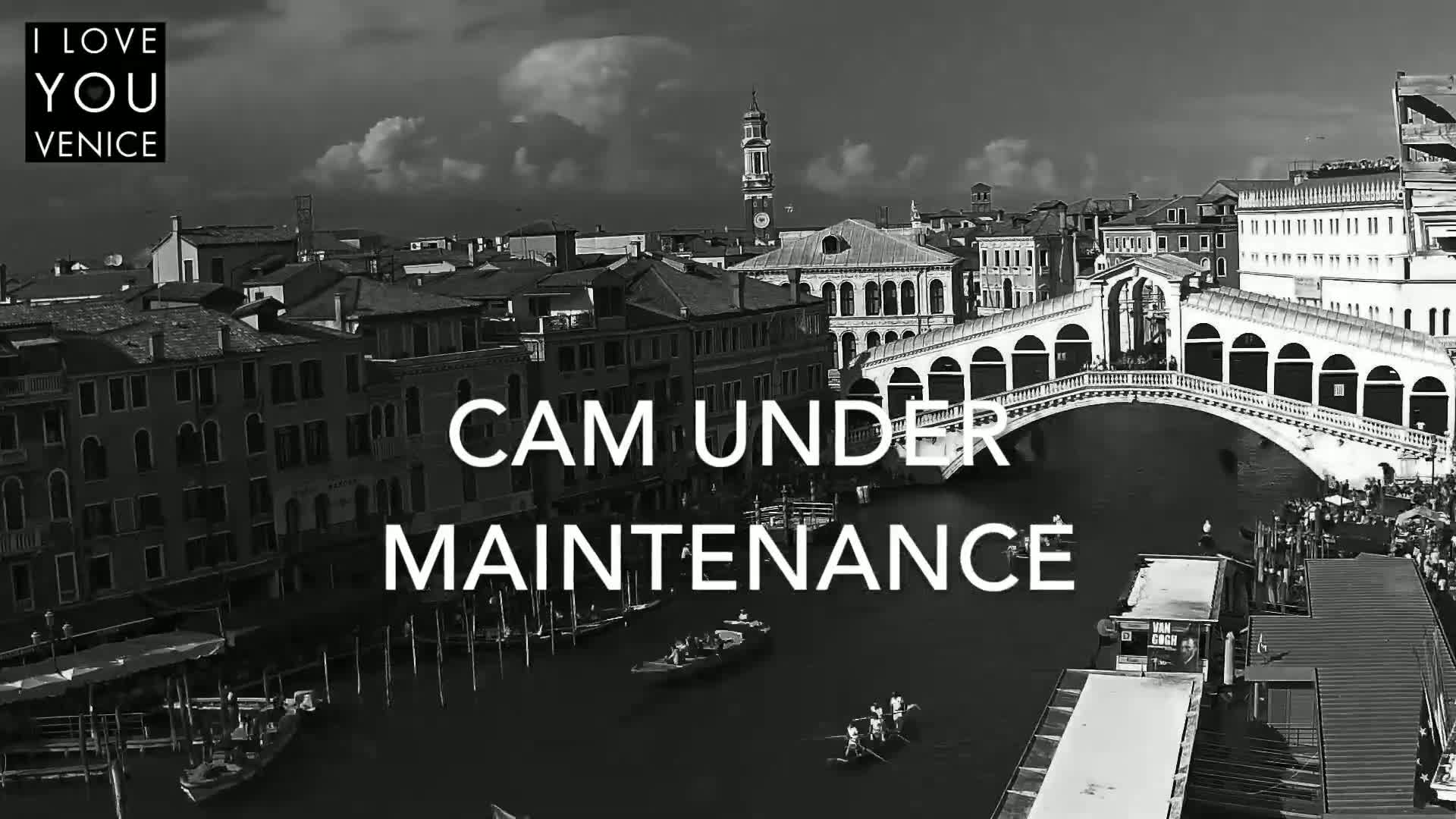A black and white, high-angle view shows a Venetian canal with gondolas, a bridge, and numerous buildings under a partly cloudy sky.
