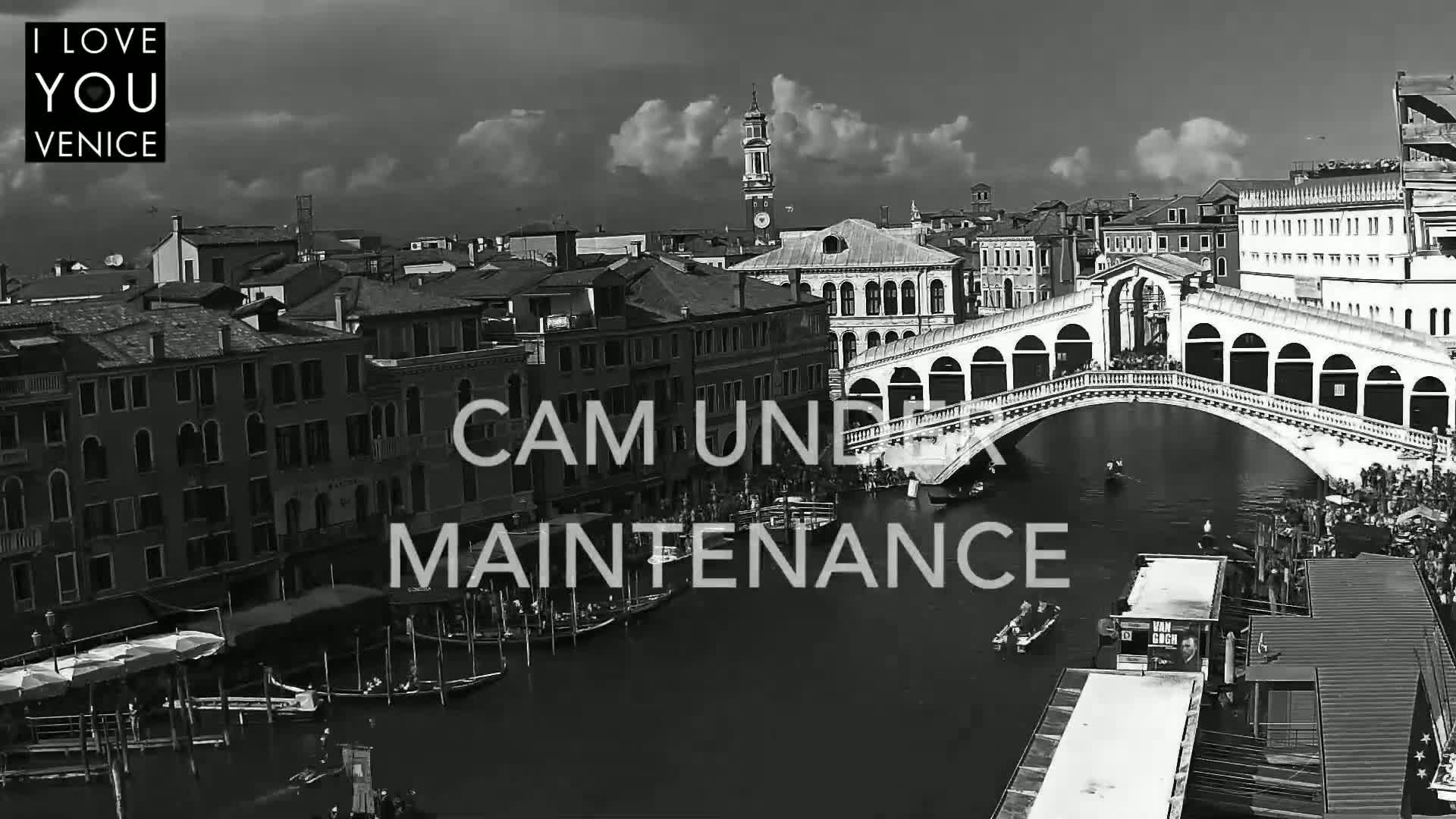A black and white, high-angle view shows a canal in Venice, Italy, with a bridge spanning the waterway and many boats and buildings lining the canal under a partly cloudy sky.
