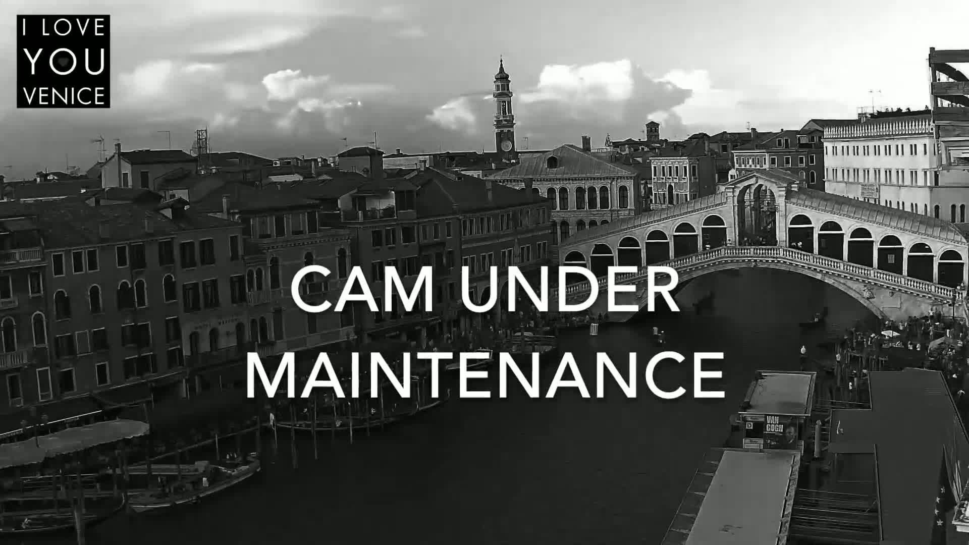 A black and white, high-angle view of a canal in Venice, Italy, shows a bridge and numerous buildings under a partly cloudy sky.