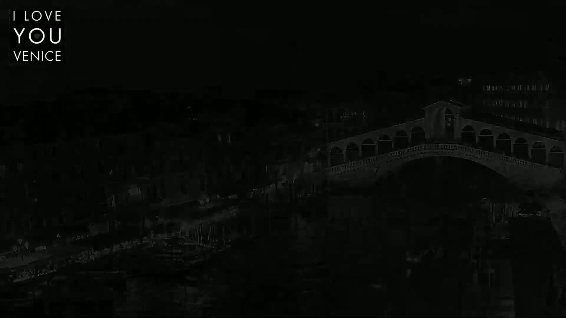 A dark, nighttime image shows a bridge over a canal, with buildings lining the waterway.