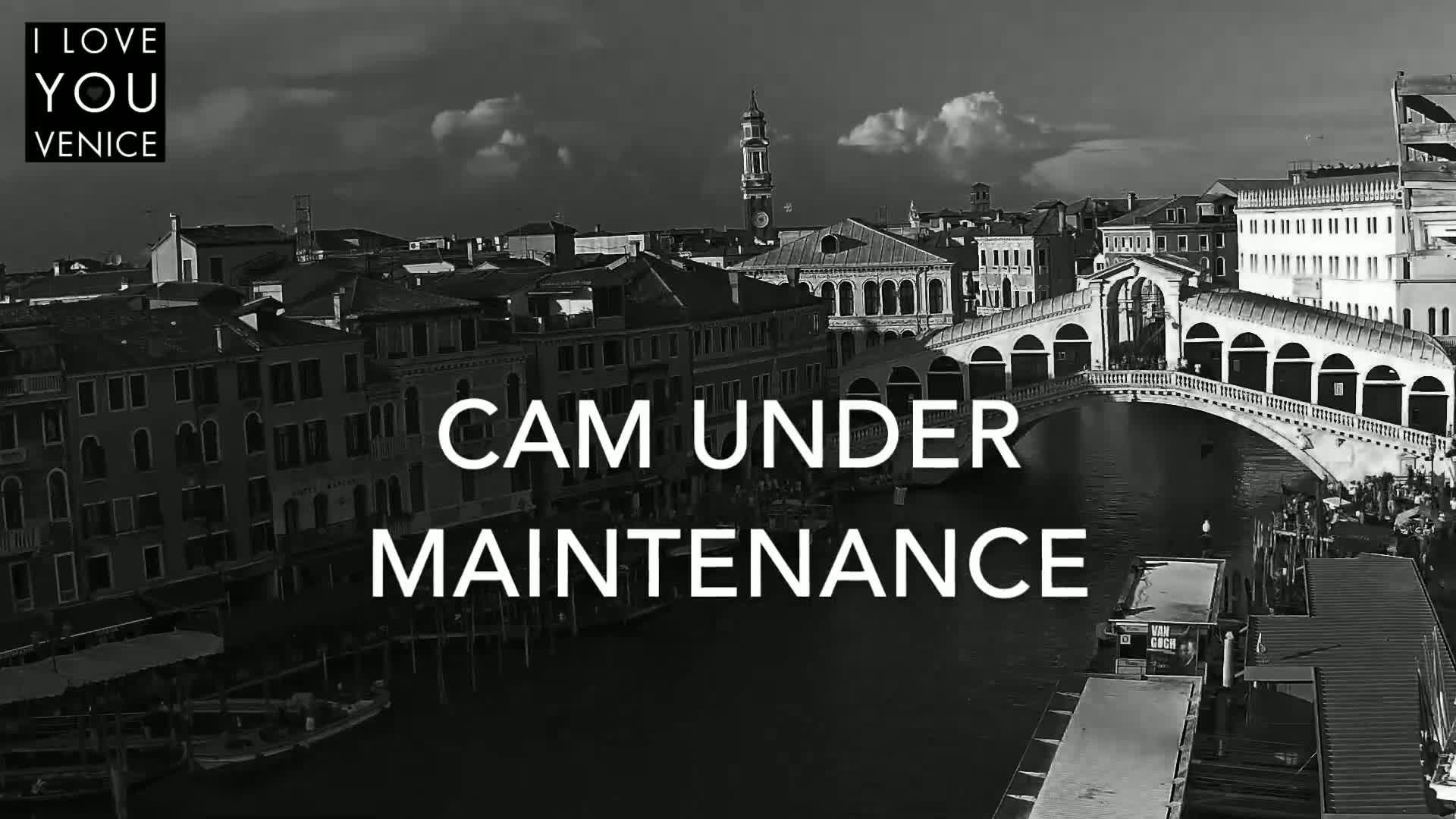 A black and white, high-angle view shows a Venetian canal with a bridge, boats, and buildings under a partly cloudy sky.