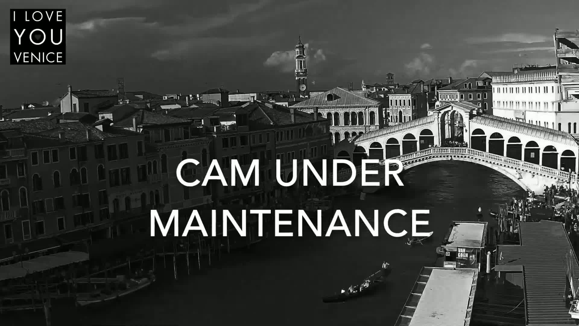 A black and white aerial view shows a canal in Venice, Italy, with a bridge and numerous buildings under a partly cloudy sky.
