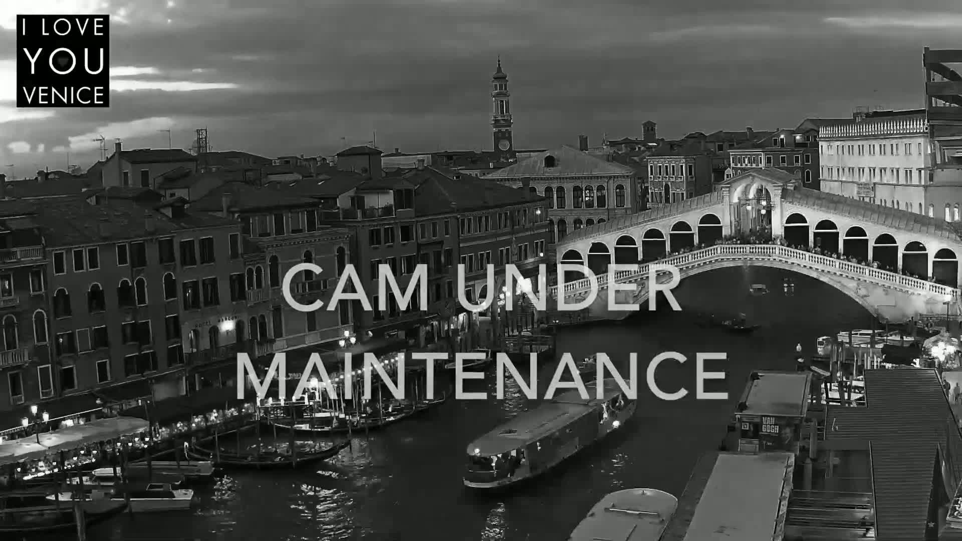 A black and white nighttime view of a Venetian canal with gondolas and a bridge, under a partly cloudy sky.