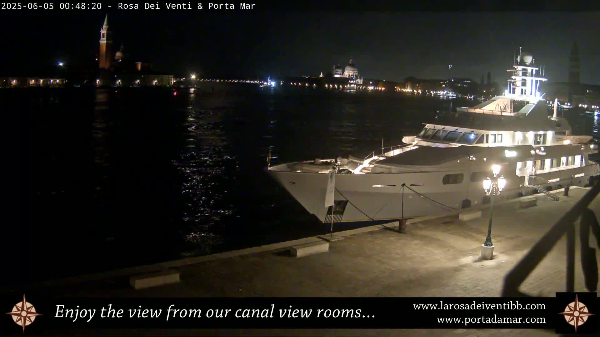 A large yacht is docked at a waterfront at night, with city lights and a bell tower visible across the water.