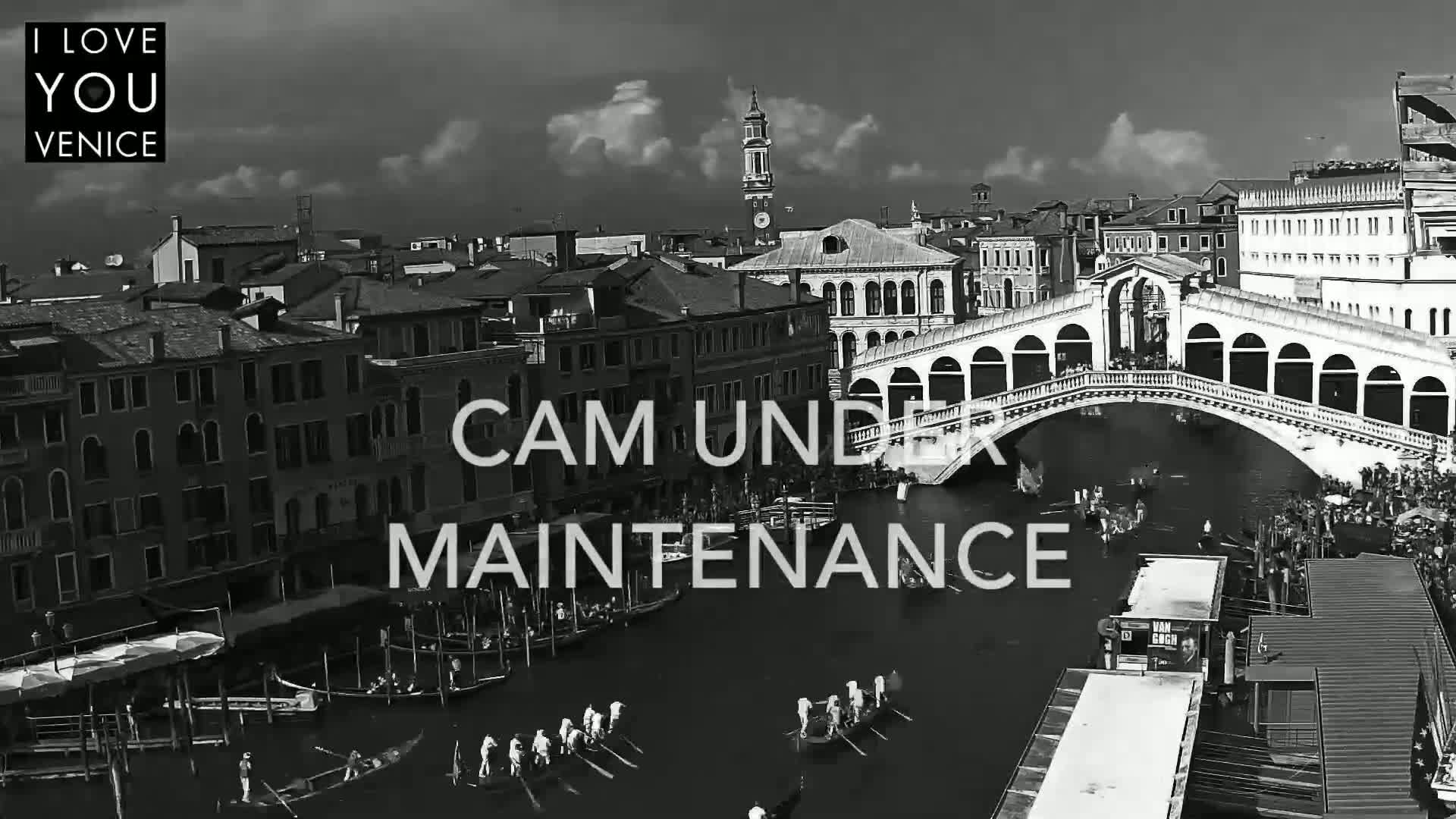 A black and white aerial view shows gondolas on a canal in Venice, Italy, under a partly cloudy sky, with the Rialto Bridge in the background.