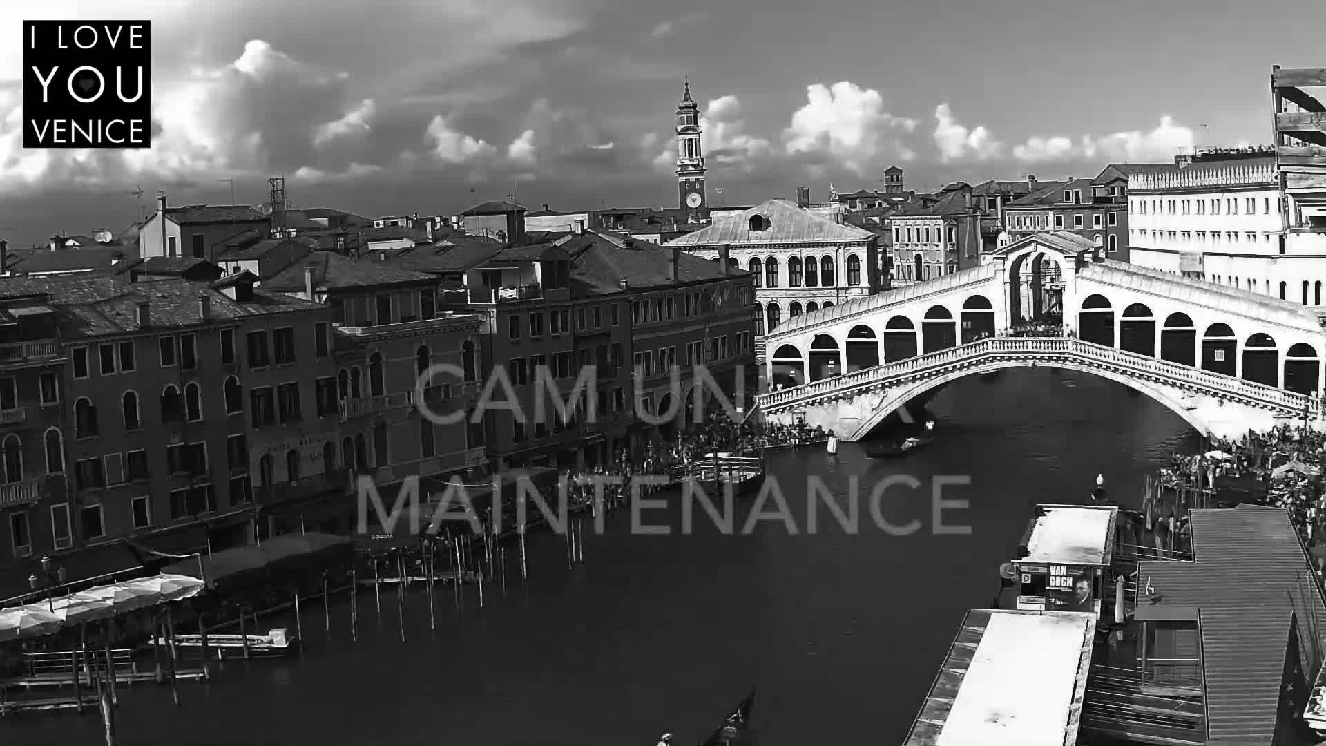 A black and white, high-angle view shows the Rialto Bridge in Venice on a partly cloudy day, with gondolas and people visible on the water and surrounding buildings.