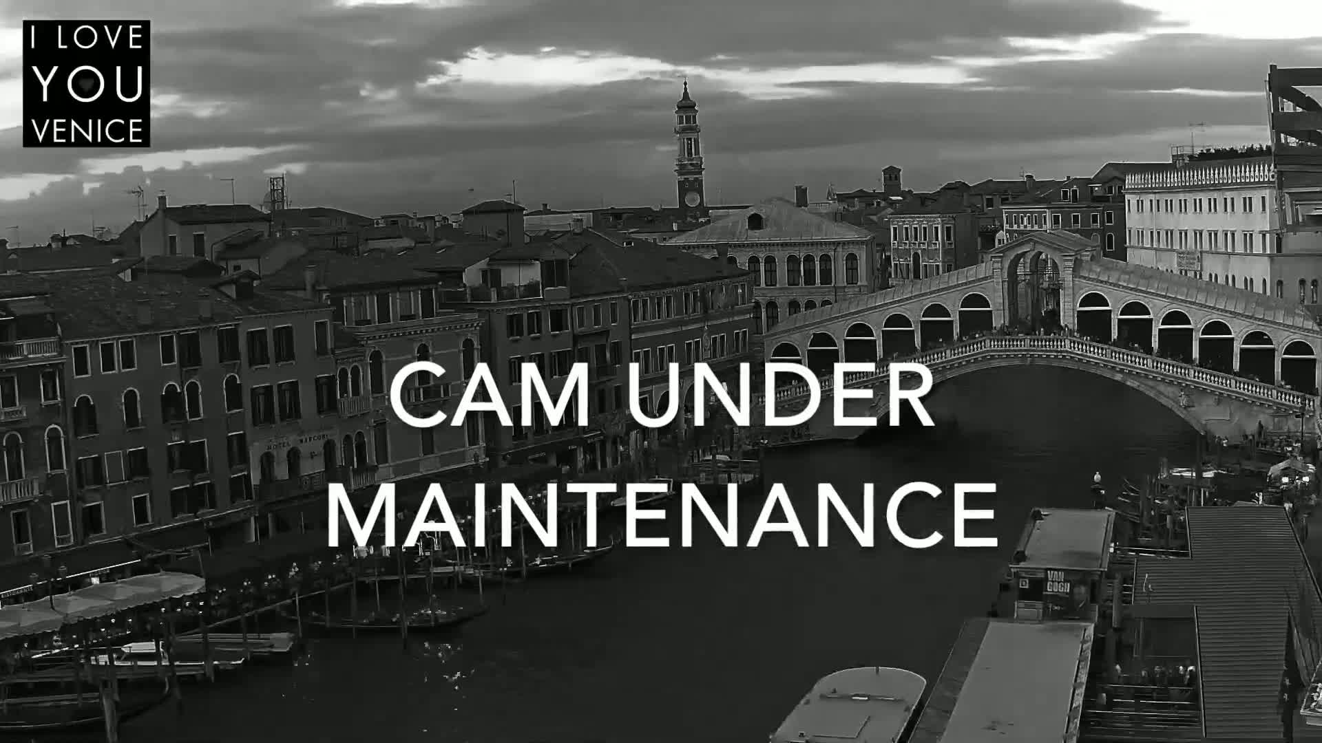 A black and white, high-angle view of a canal in Venice, Italy, shows buildings lining the waterway, with a bridge arching over the canal under a cloudy sky.