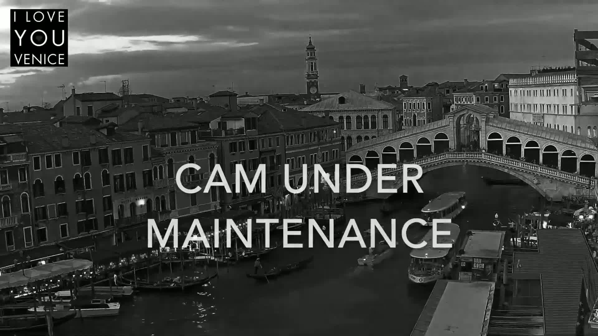 A black and white, nighttime view of a canal in Venice, Italy, shows gondolas and other boats moored along the banks, with the Rialto Bridge visible in the background under a partly cloudy sky.