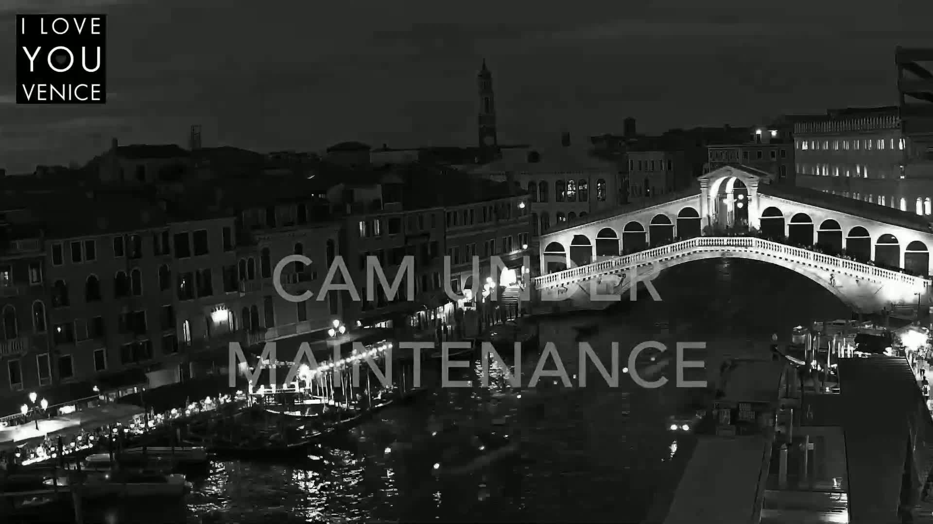 A black and white nighttime view of a bridge over a canal in Venice, with boats and buildings lining the waterway.