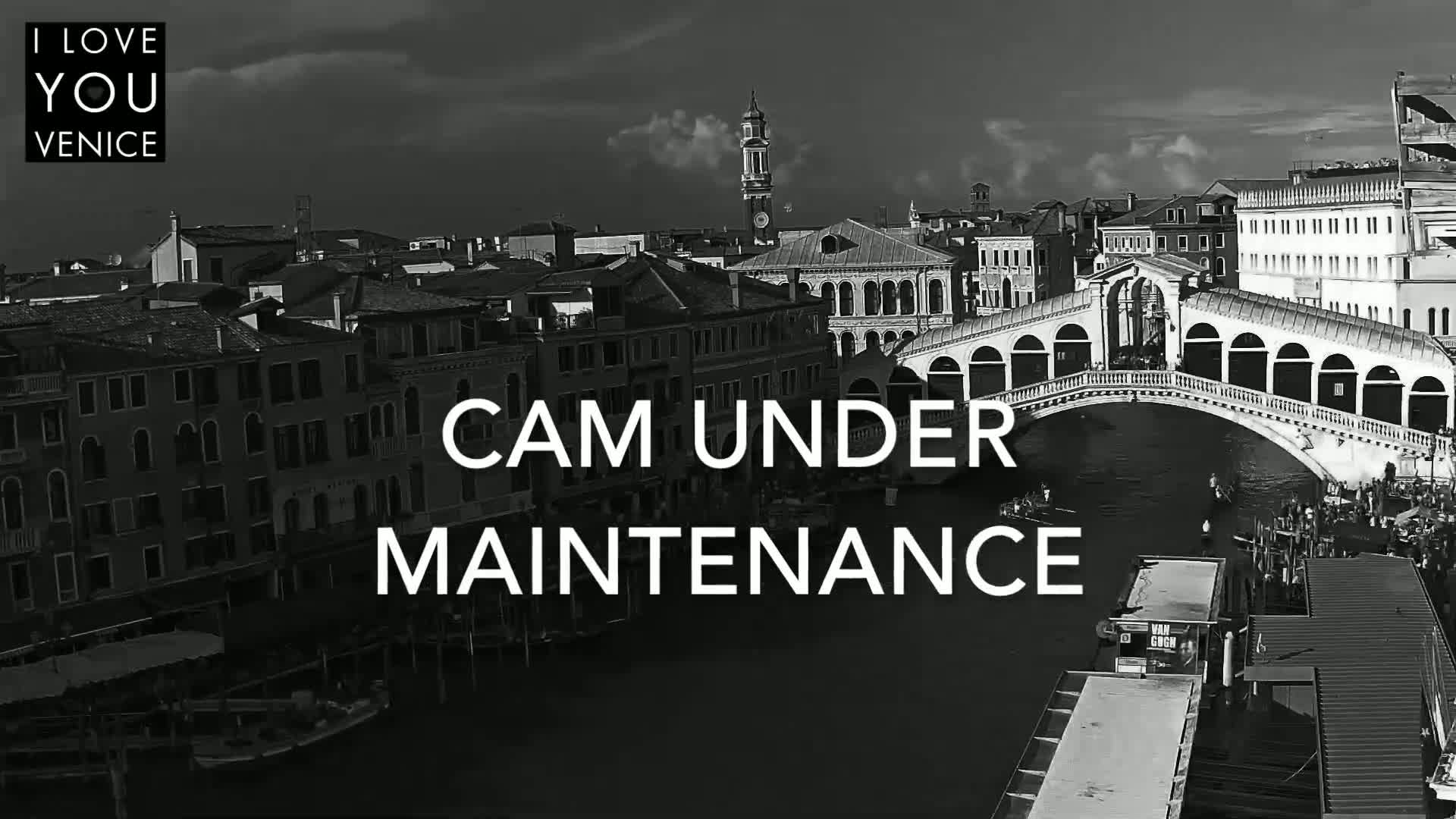 A black and white, high-angle view shows a canal in Venice, Italy, with a bridge spanning the waterway, under a partly cloudy sky.