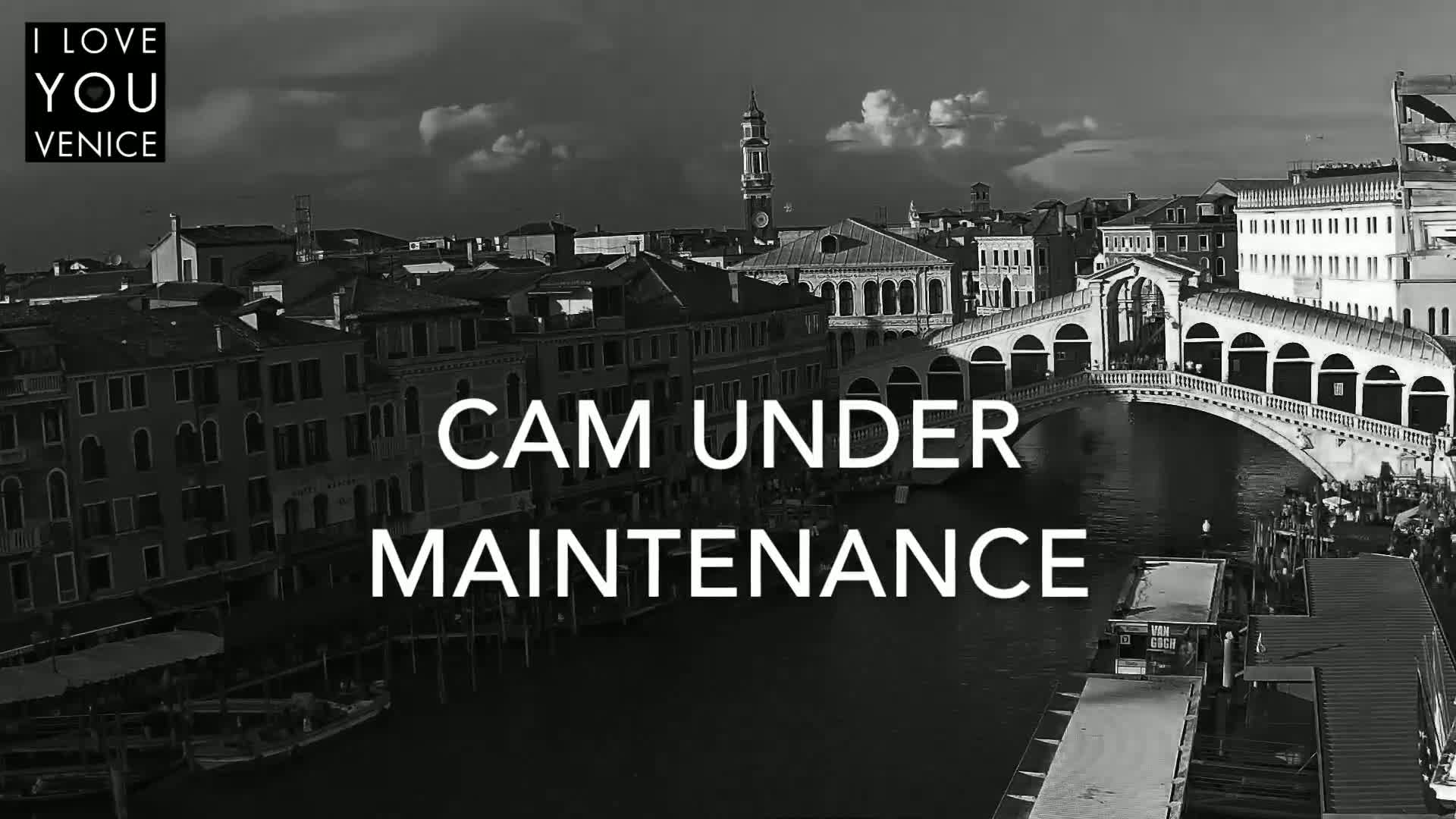 A black and white, high-angle view shows a canal in Venice with a bridge, buildings, and boats under a partly cloudy sky.