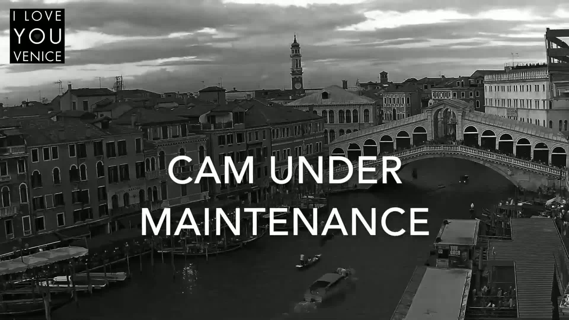 A black and white aerial view of a Venetian canal shows buildings, a bridge with people on it, boats, and overcast skies.